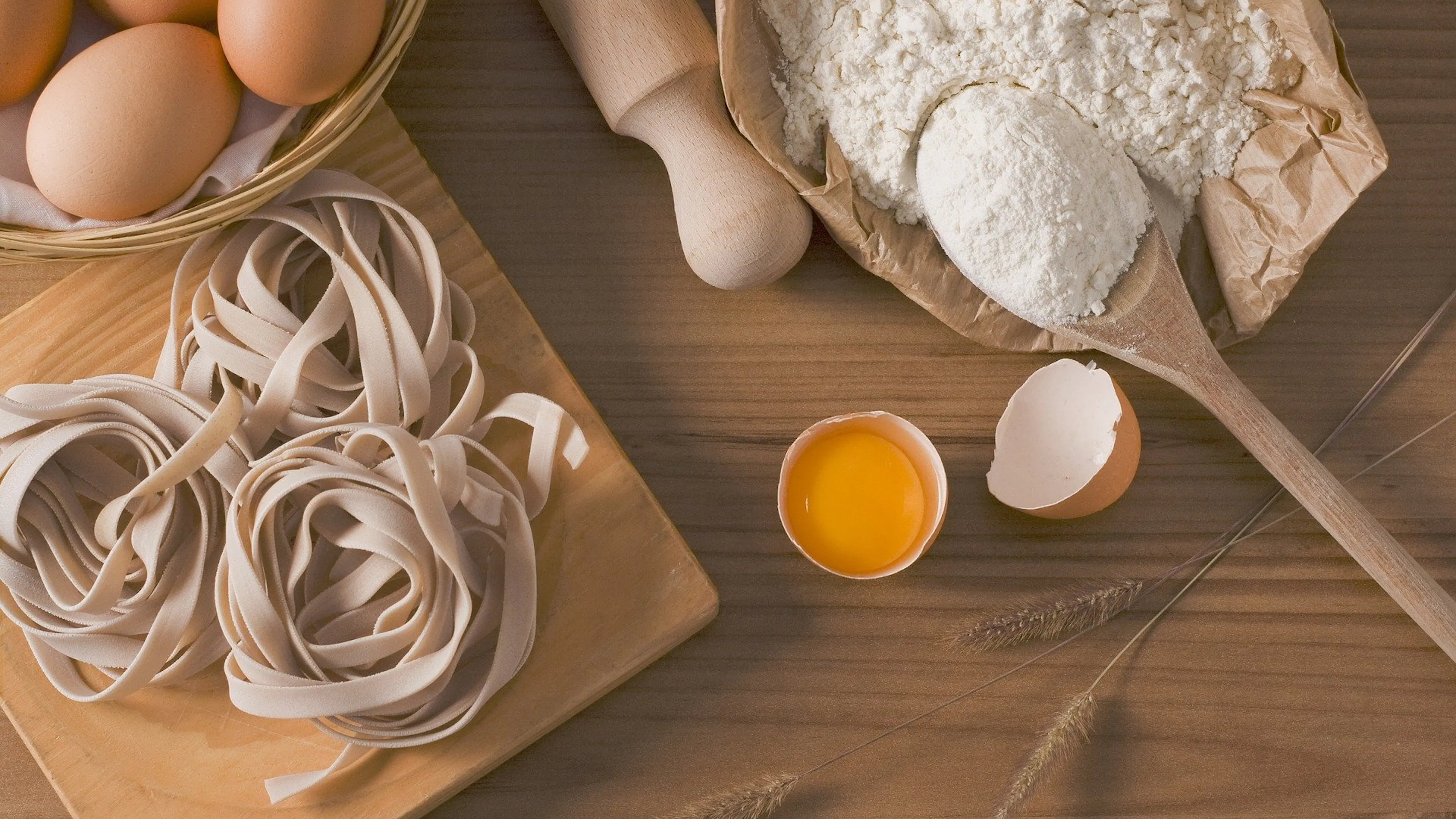 Eggs flour and wooden spoons next to freshly made fettuccini pasta on a wooden table seen from above