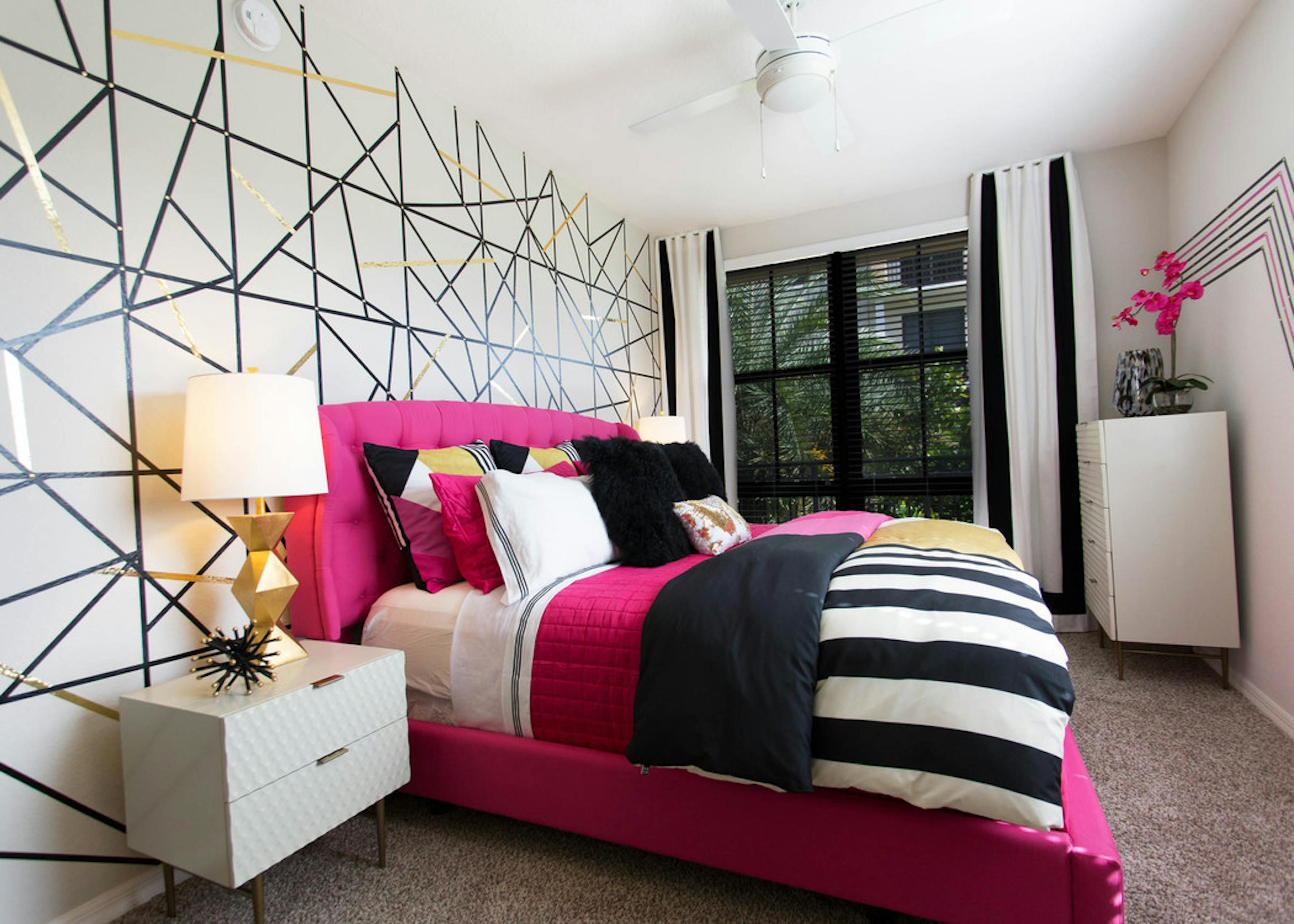 AMLI Dadeland master bedroom with geometric accent wall, modern table lamp, magenta and black bedding and a window view