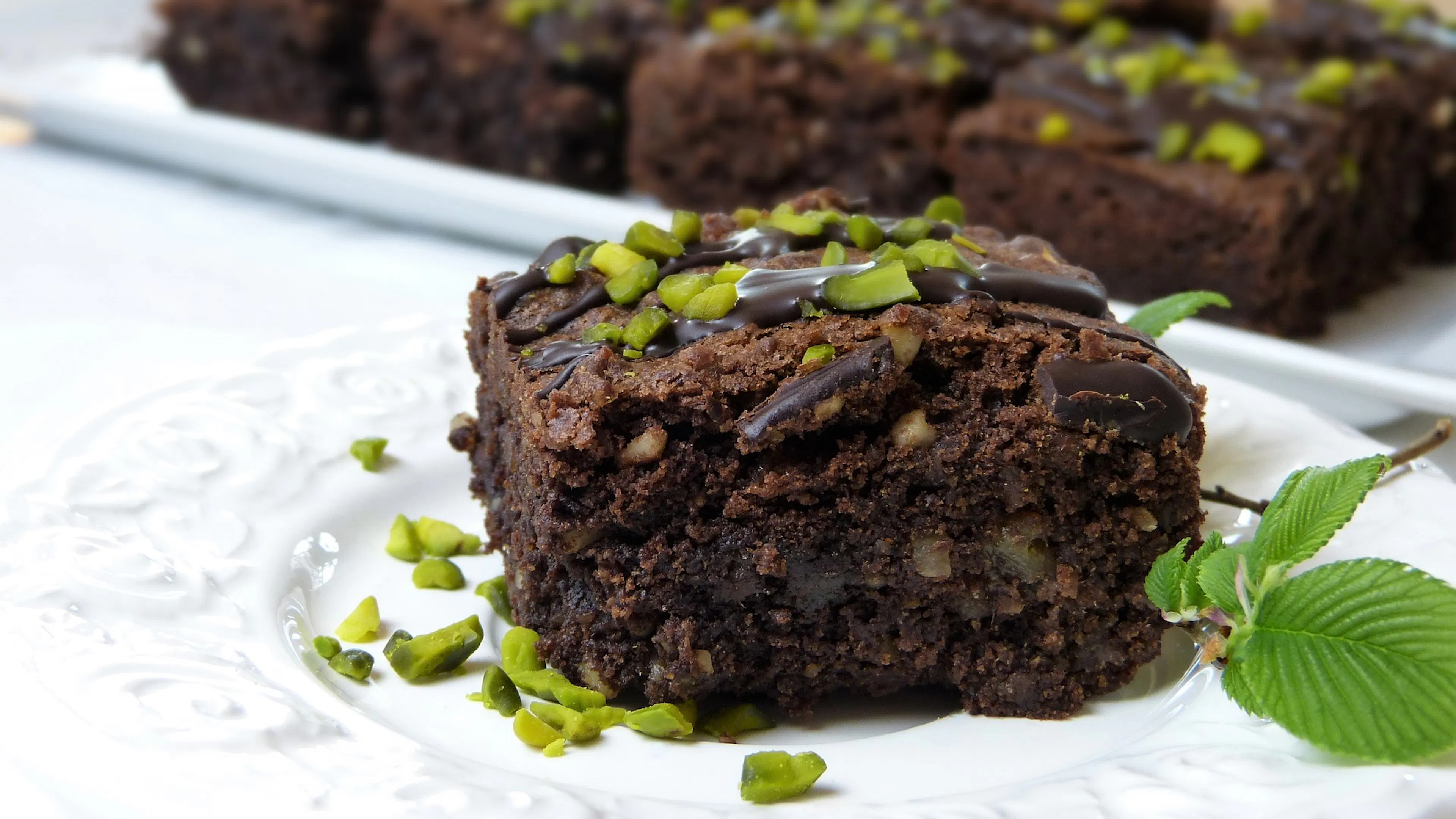 A chocolate brownie resting on a white plate and topped with crushed pistachios and a sprig of fresh mint leaves