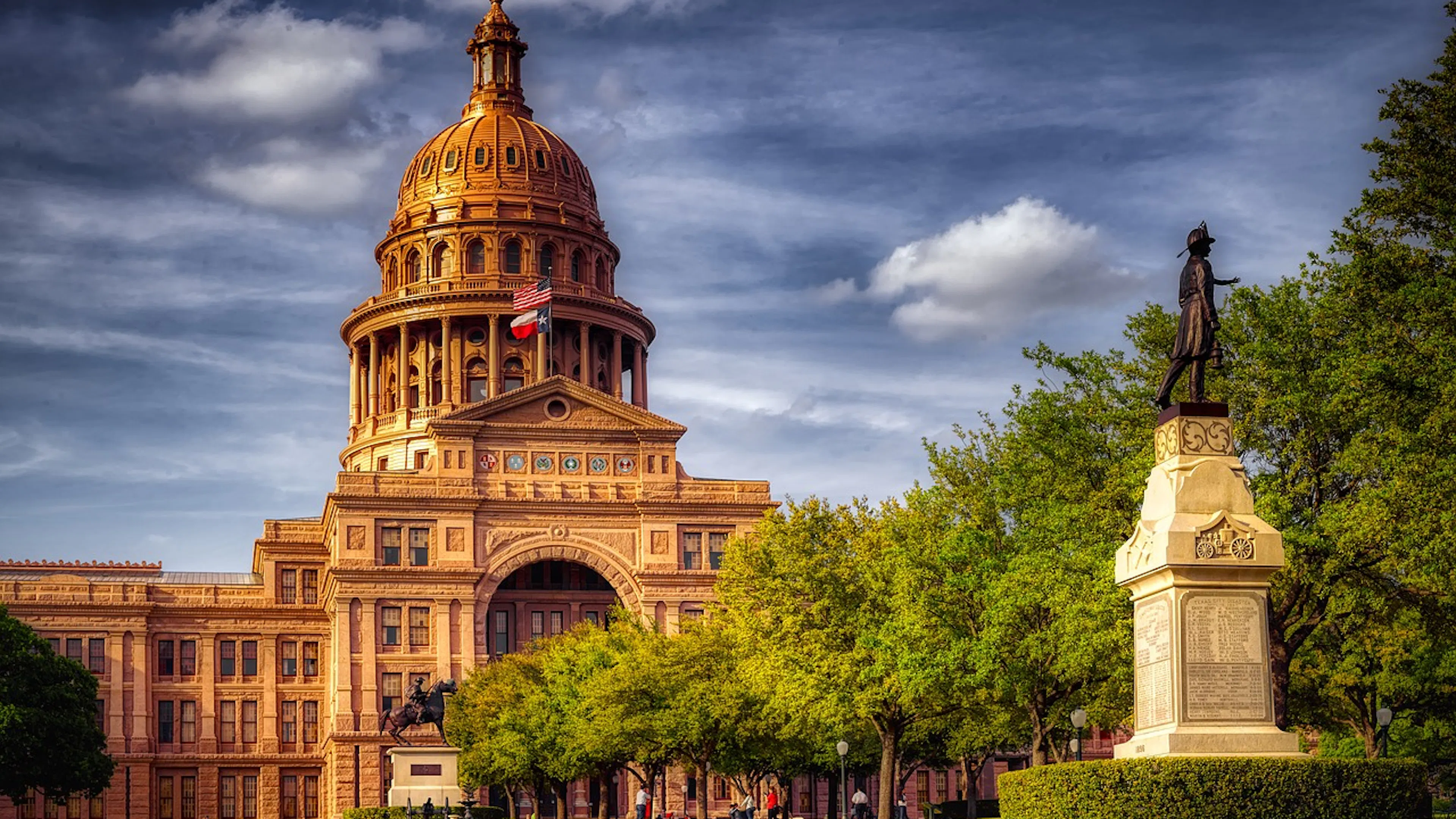 The intricately built Texas State Capitol building lit up during a cloudy sunset with trees around a monument statue