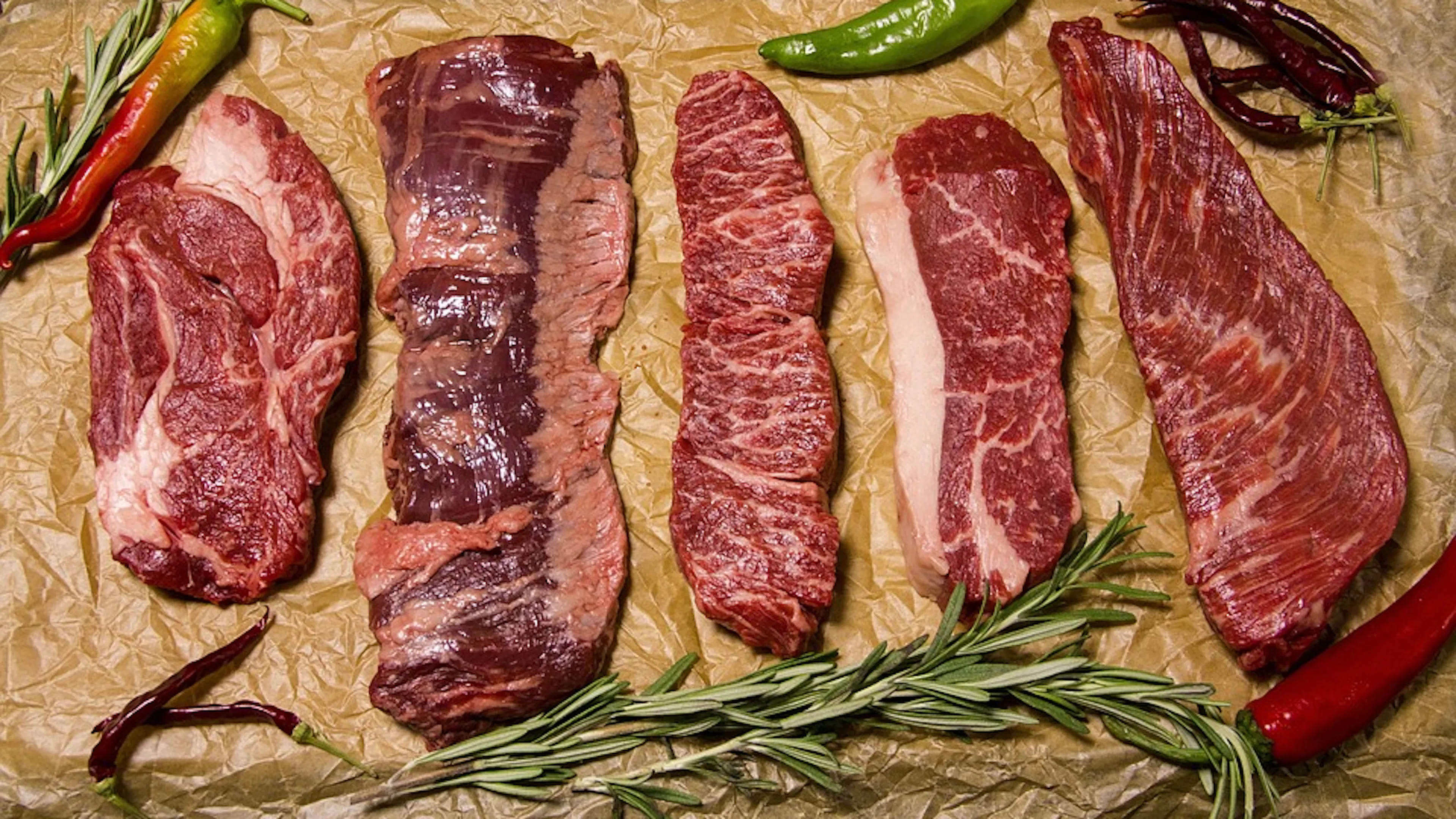 Five different cuts of beef steak lying beside each other on brown paper surrounded by fresh herbs