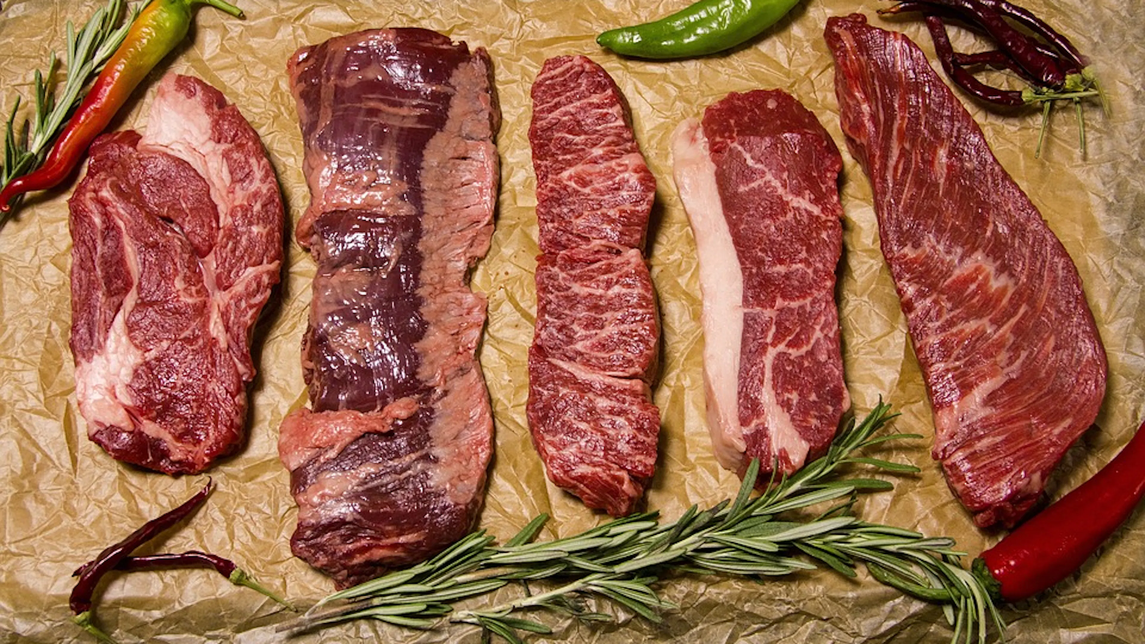 Five different cuts of beef steak lying beside each other on brown paper surrounded by fresh herbs