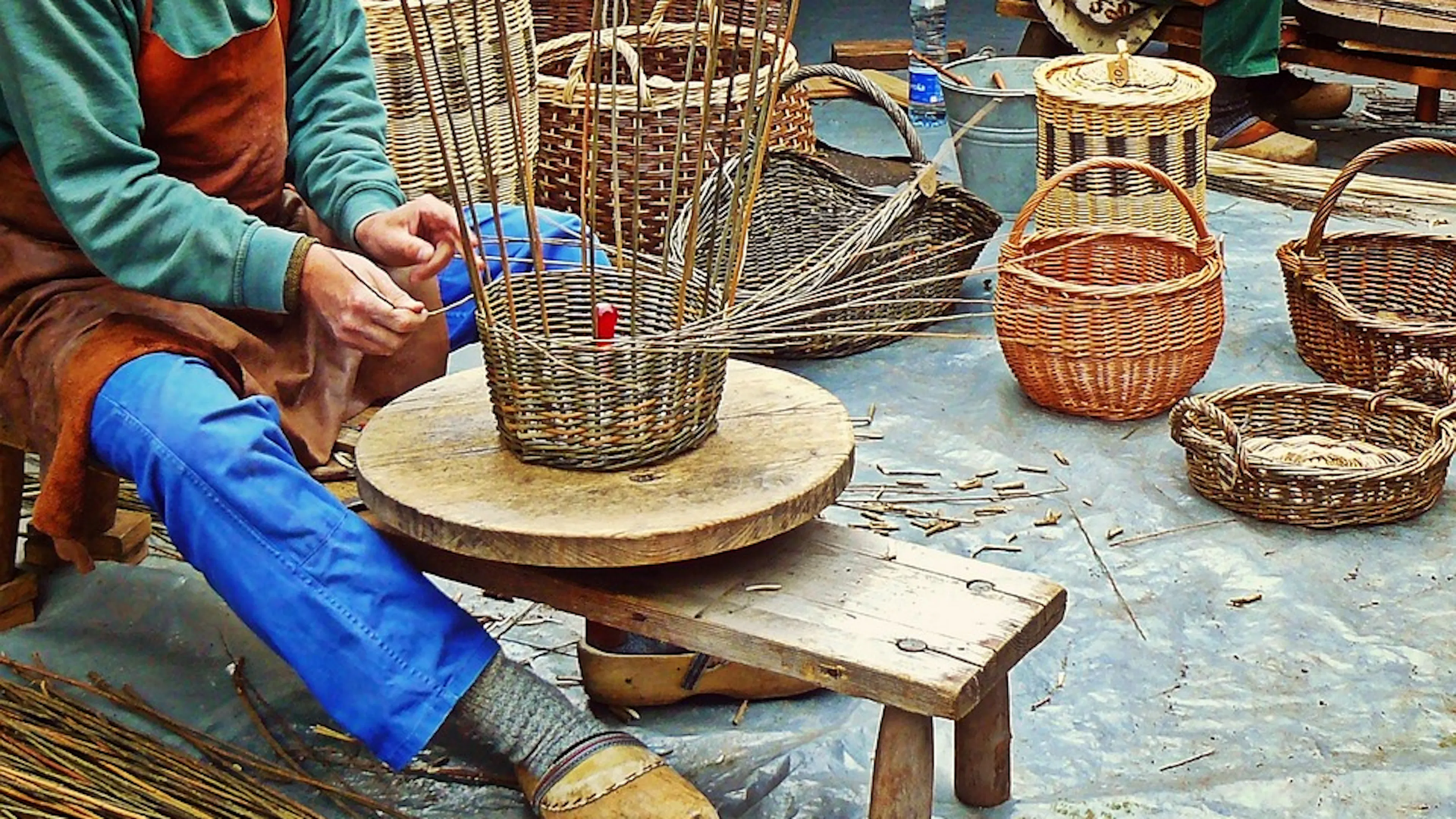 A person seated on a short stool outdoors weaving a tall wicker basket