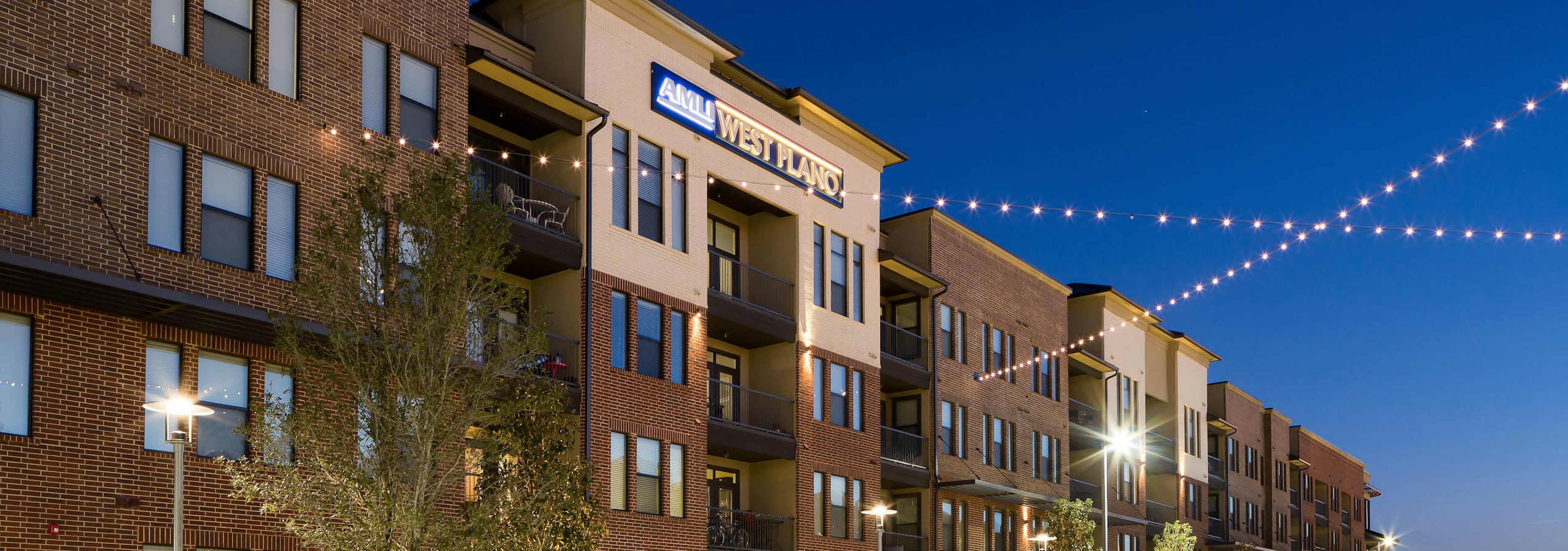 Exterior of AMLI West Plano apartment community at dusk with trees and string lights in front of the building facade