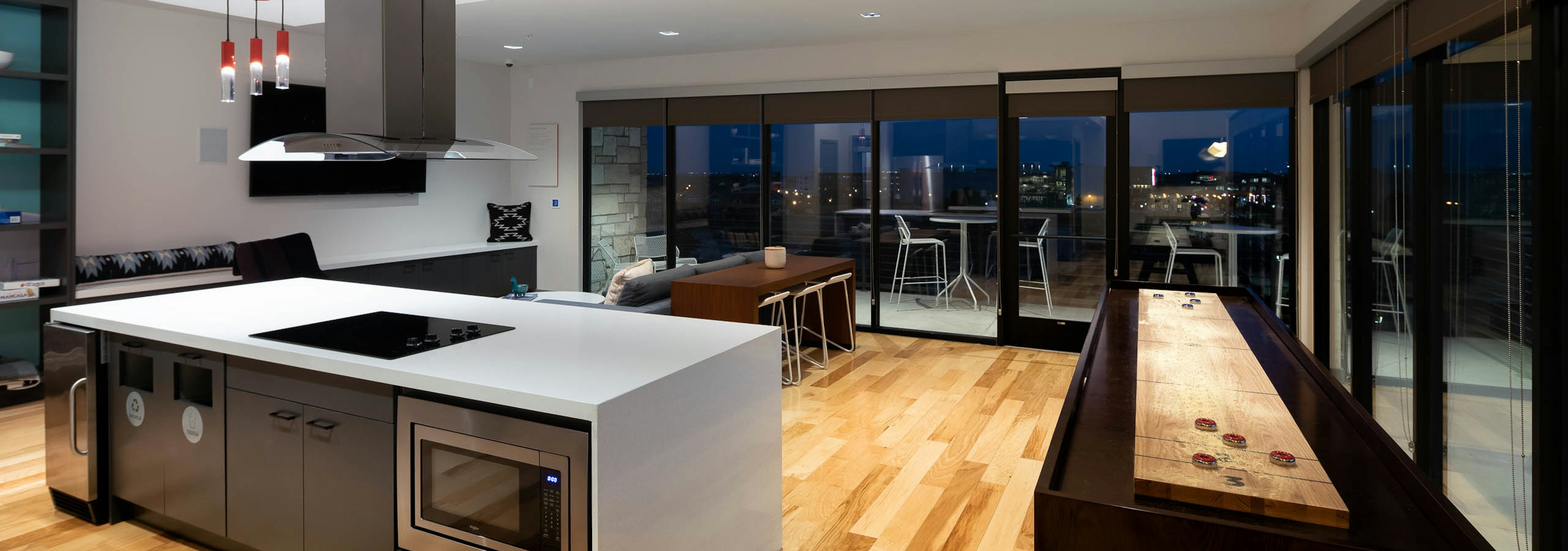 Clubroom at AMLI on Aldrich with floor to ceiling windows showing nighttime view with light wood floors and spacious kitchen