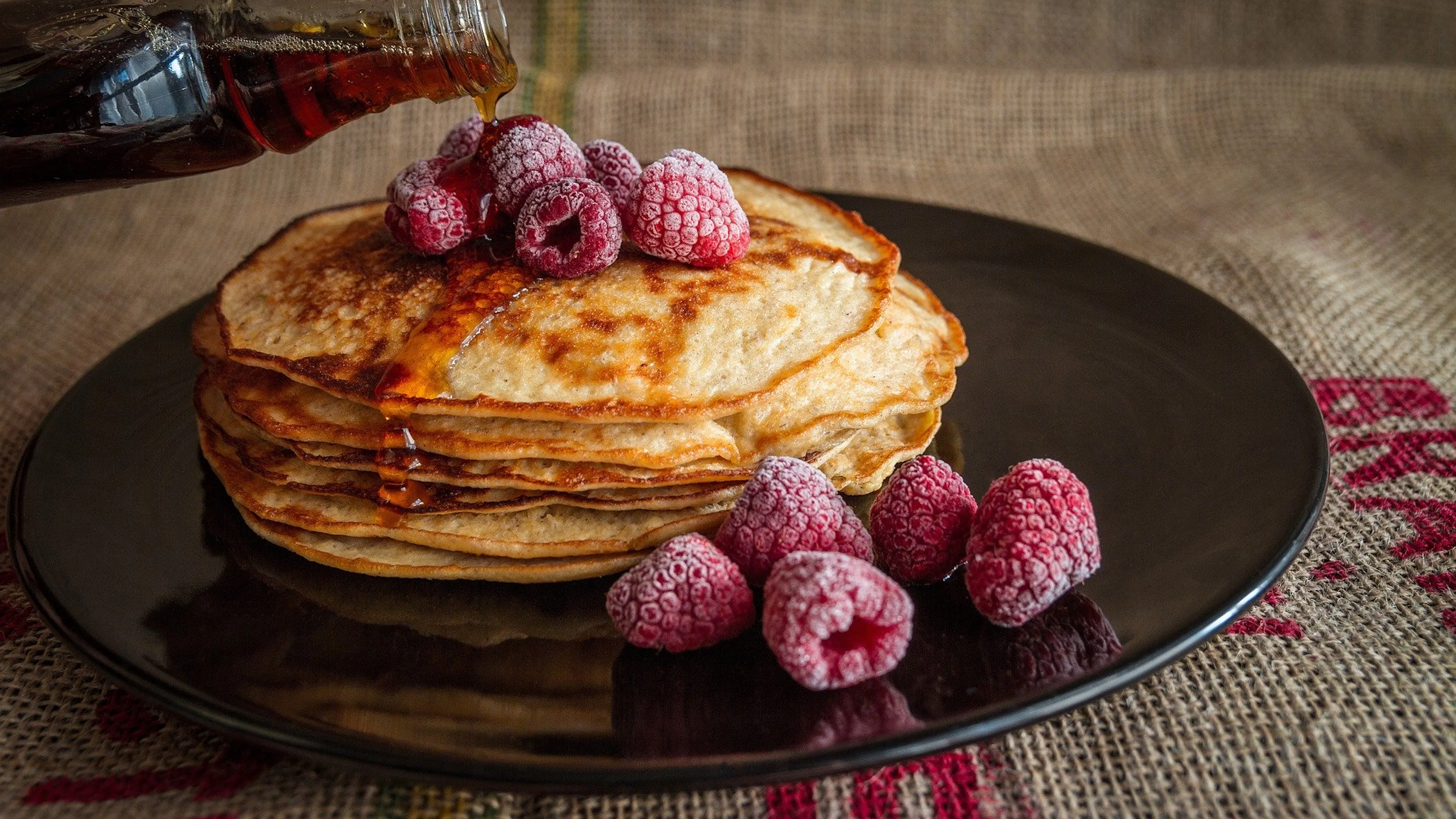 A stack of 6 thin pancakes on a black plate with frozen raspberries sprinkled on top and a bottle pouring syrup