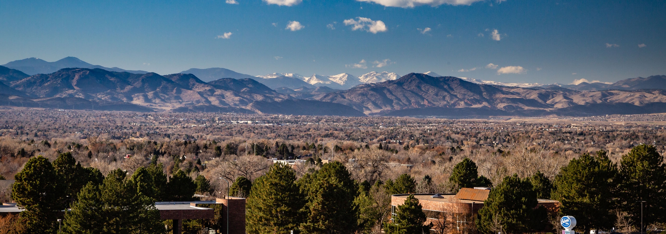 Daytime aerial view above AMLI Littleton Village apartments with the Rocky Mountains from afar, blue sky and lush greenery