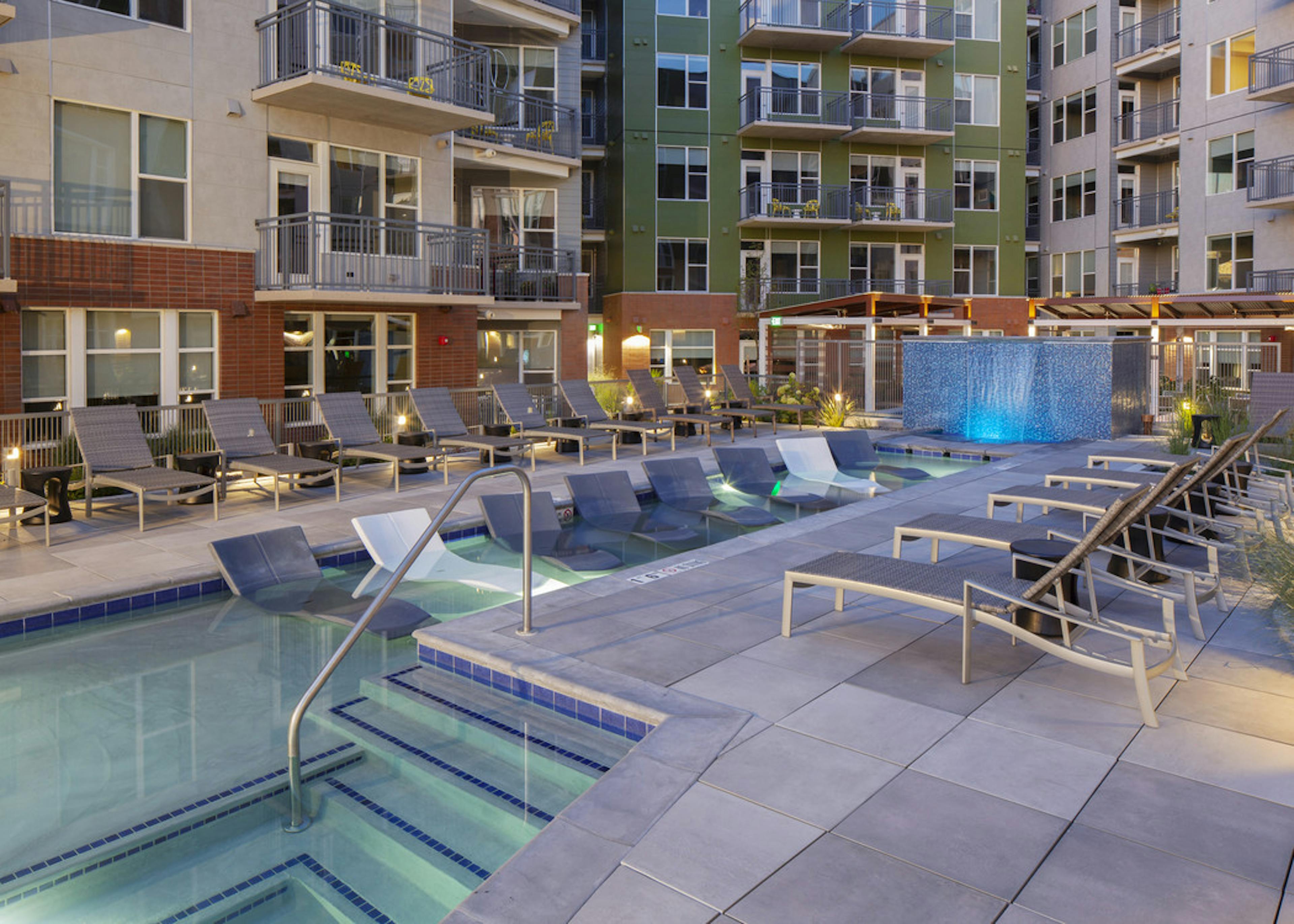 A night time view of the pool at AMLI Riverfront Green apartments with several lounge chairs and a tile waterfall 