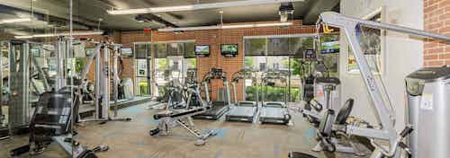 Fitness center at AMLI Las Colinas apartments with cardio and weight machines against a red brick wall with mounted TVs