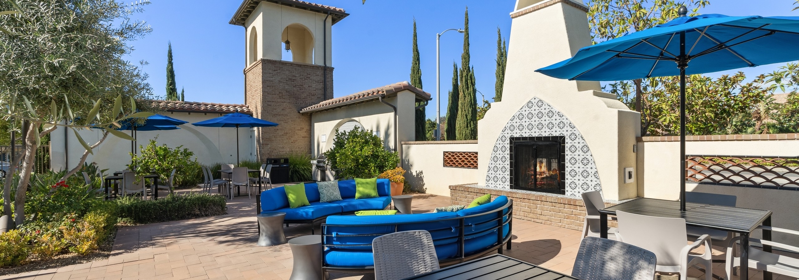 Daytime view of outdoor lounge with blue cushioned seating and umbrellas next to fireplace at AMI Spanish Hills apartments