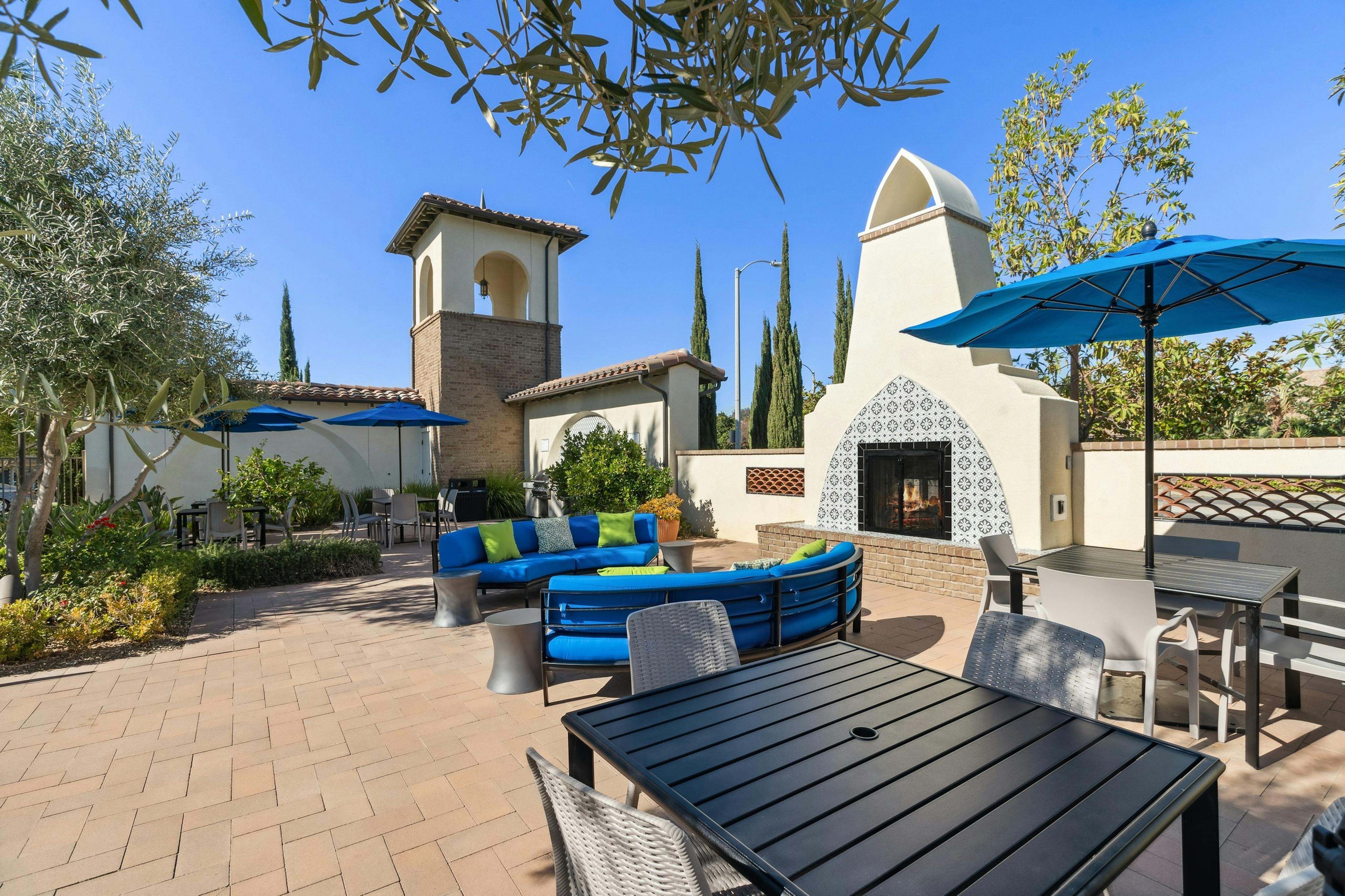 Daytime view of outdoor lounge with blue cushioned seating and umbrellas next to fireplace at AMI Spanish Hills apartments