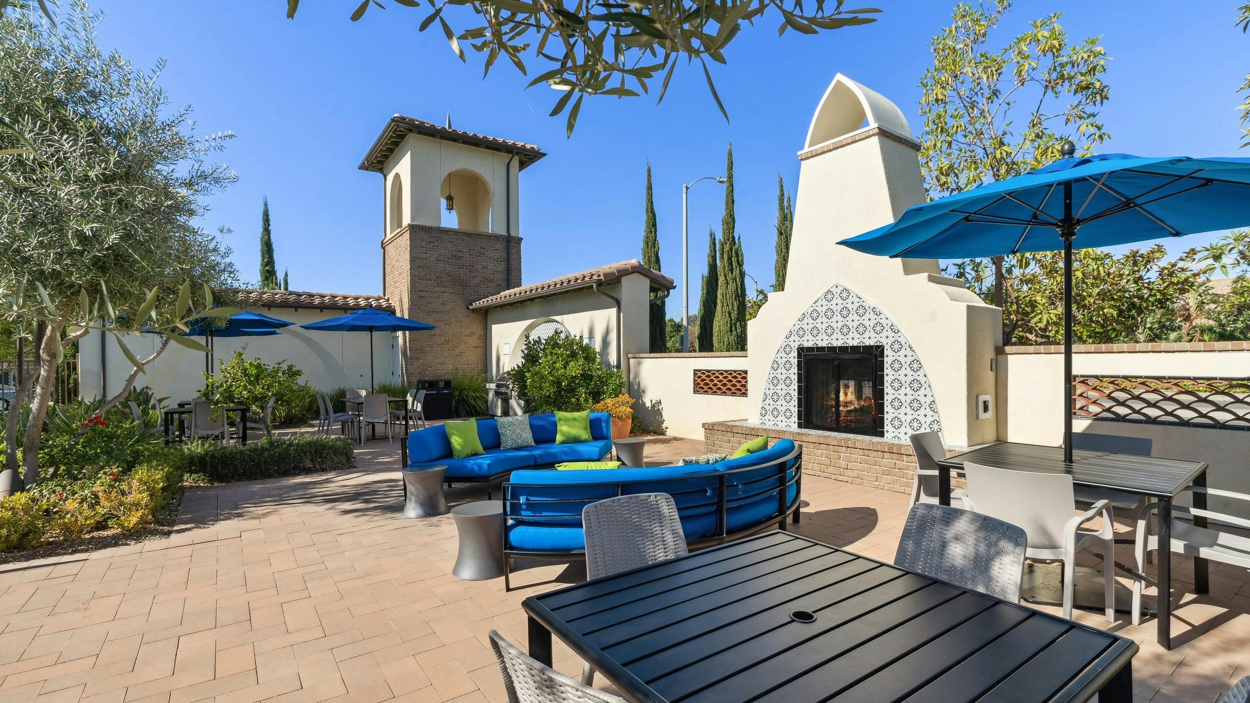 Daytime view of outdoor lounge with blue cushioned seating and umbrellas next to fireplace at AMI Spanish Hills apartments