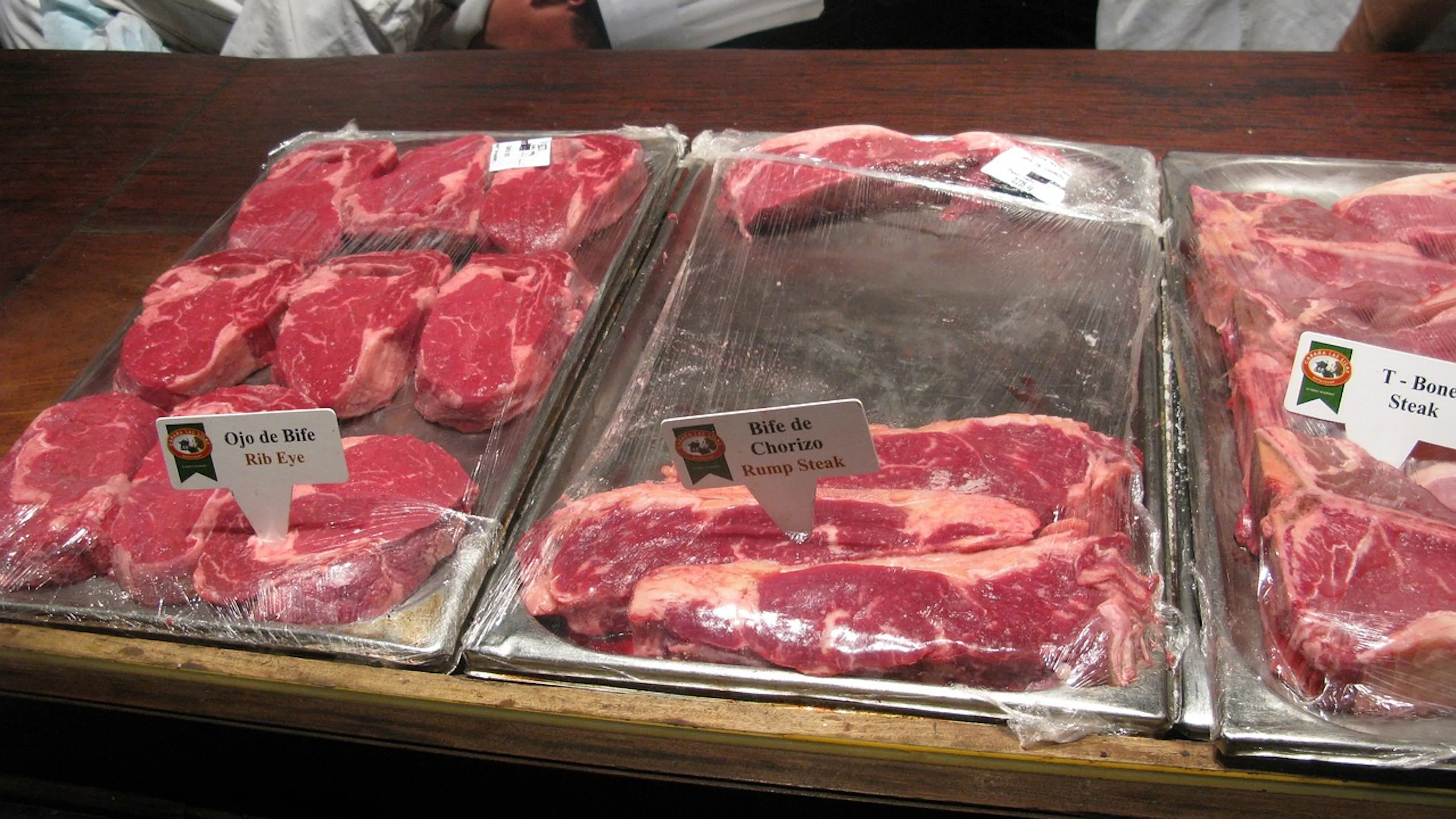 Three metal trays with different cuts of steak on them covered with cling wrap