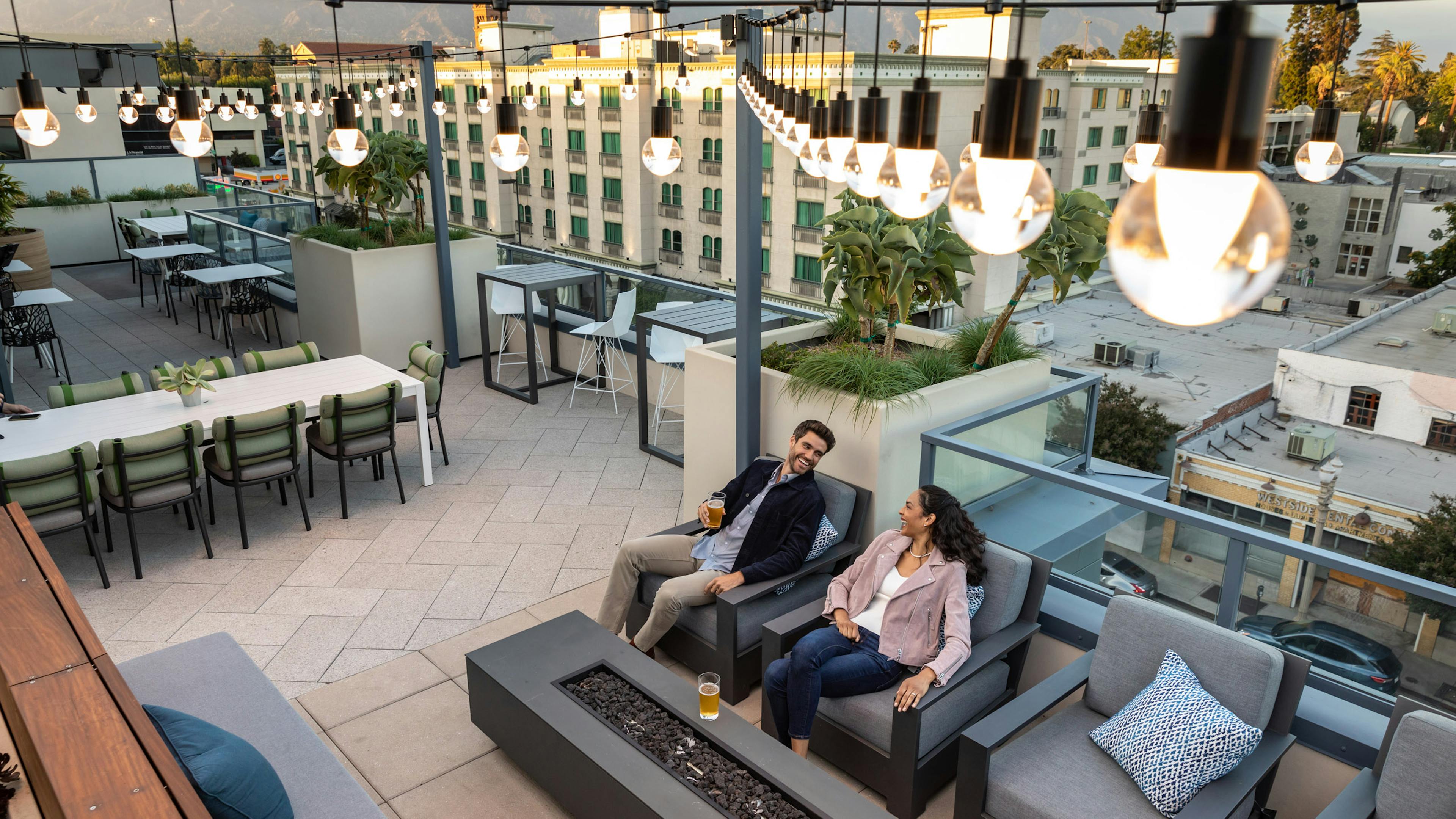 A couple sitting by the firepit and enjoying a beer under lit string lights at the rooftop sky deck at AMLI Old Pasadena
