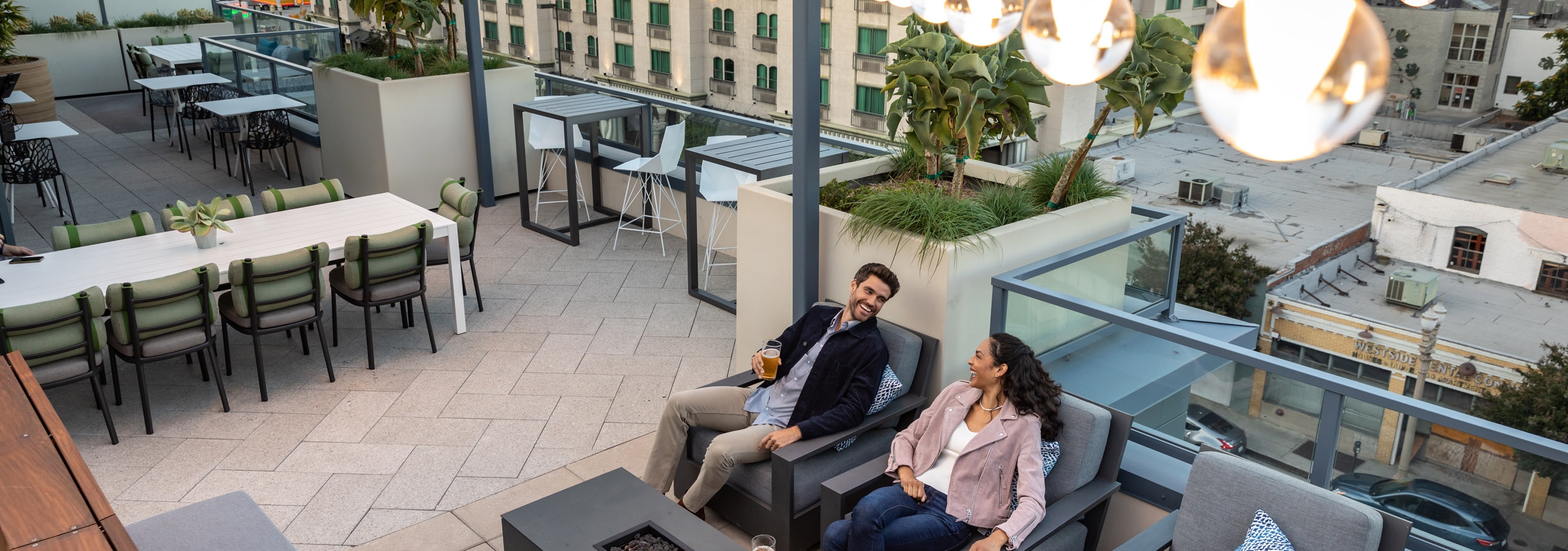 A couple sitting by the firepit and enjoying a beer under lit string lights at the rooftop sky deck at AMLI Old Pasadena