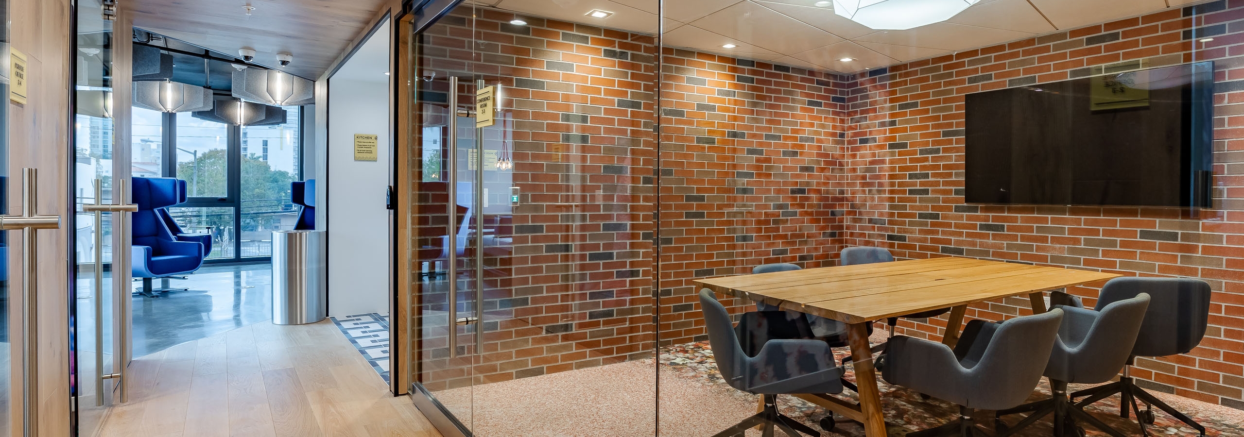 Glass enclosed conference room at AMLI Midtown Miami with wood table and 6 chairs and TV on and brick wall and adjacent hallway