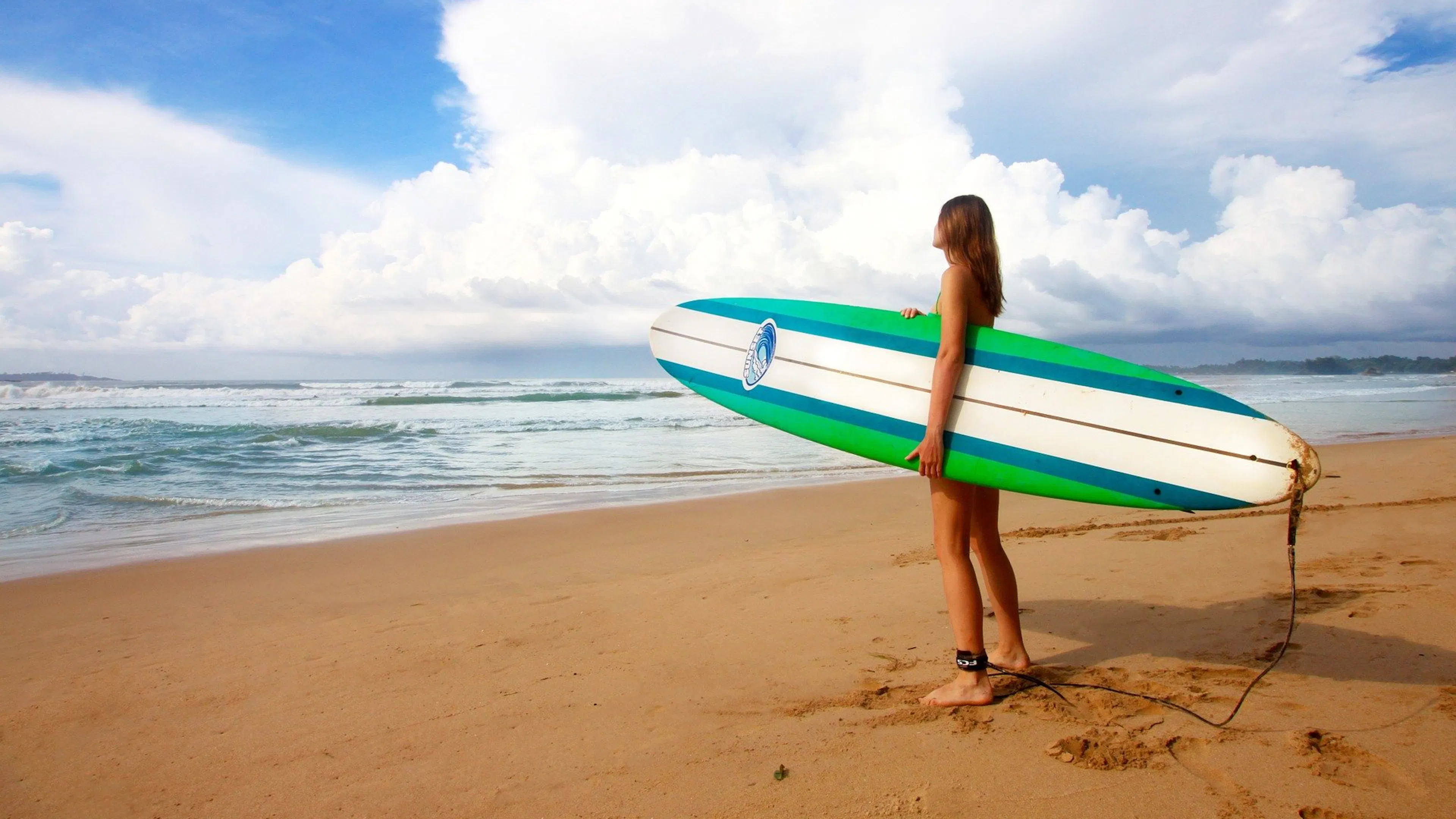 A woman holding a striped surfboard staring out into the ocean from a sandy beach