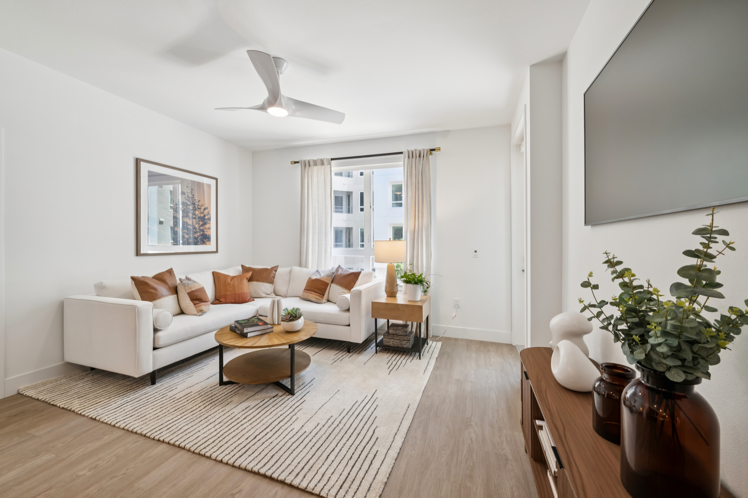 Open living room and kitchen with white sofa, round coffee table, wood cabinets, island seating, pendant lights, framed art, and plants.