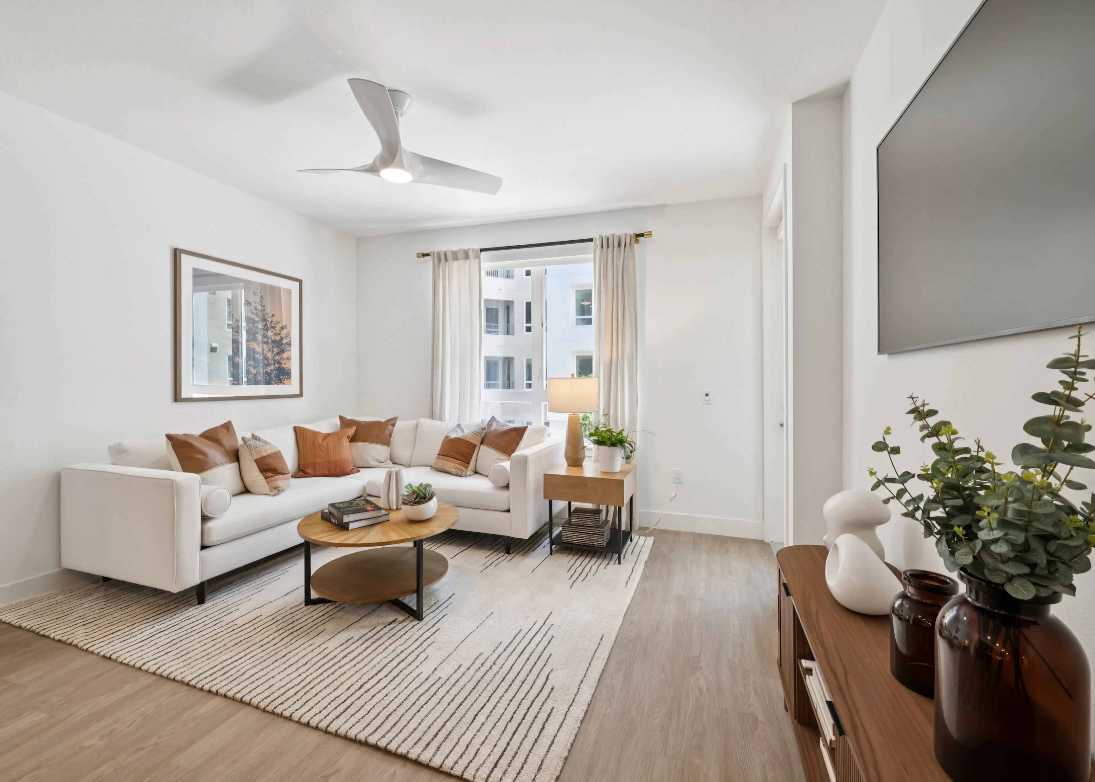 Open living room and kitchen with white sofa, round coffee table, wood cabinets, island seating, pendant lights, framed art, and plants.
