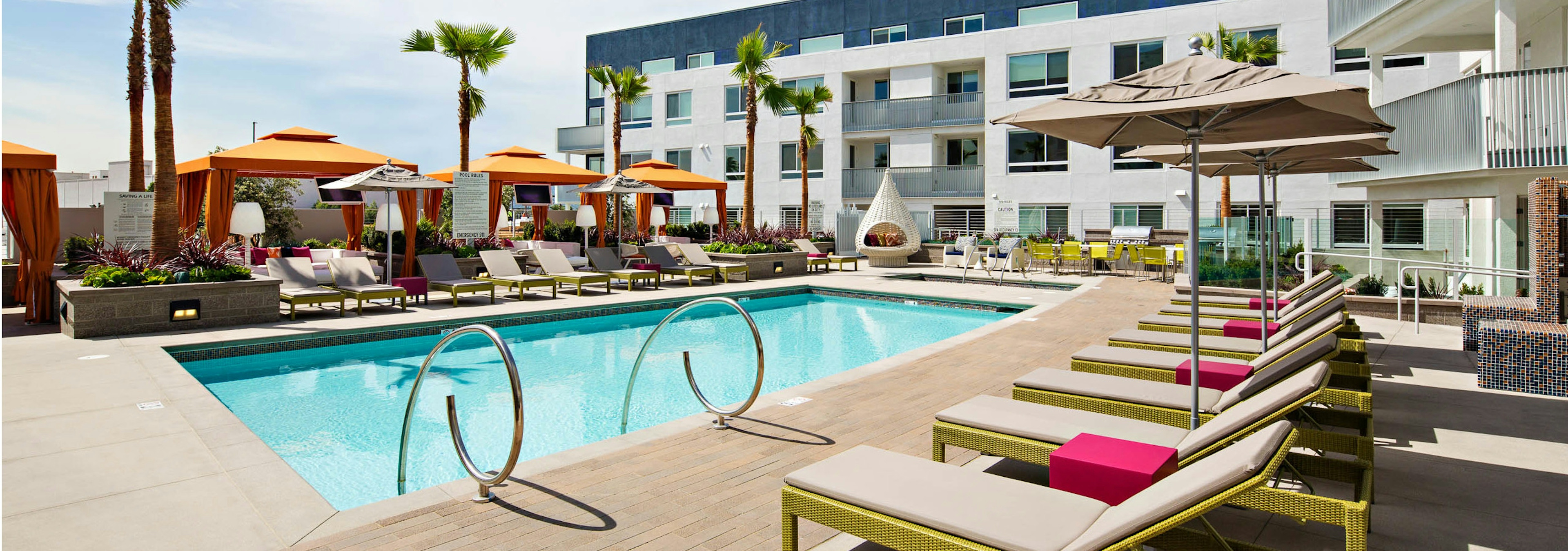 Daytime view of AMLI Lex on Orange swimming pool area with lounge seating, three orange cabanas and palm trees and blue sky