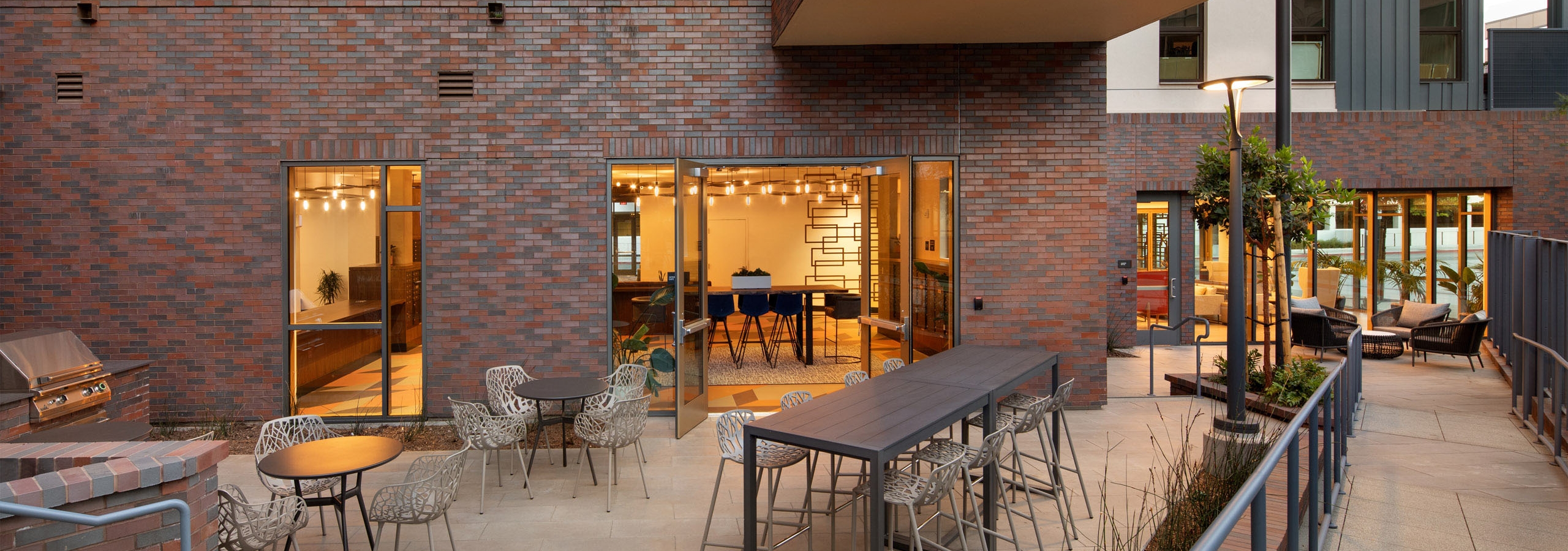 Mail lounge patio with BBQ grill and seating arrangements looking into lit up mail room at AMLI Old Pasadena