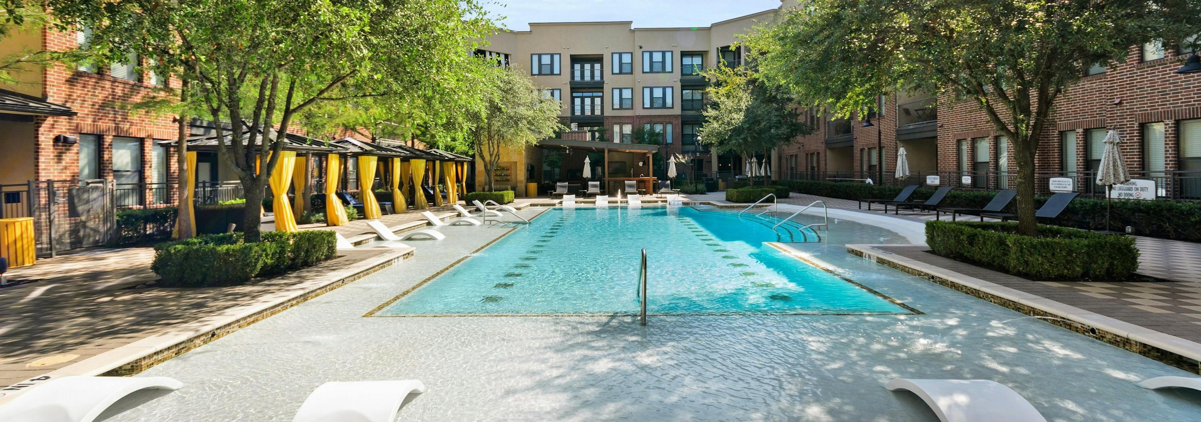Courtyard pool at AMLI West Plano apartments featuring in water seating and surrounded by lush landscaping and lounge seating