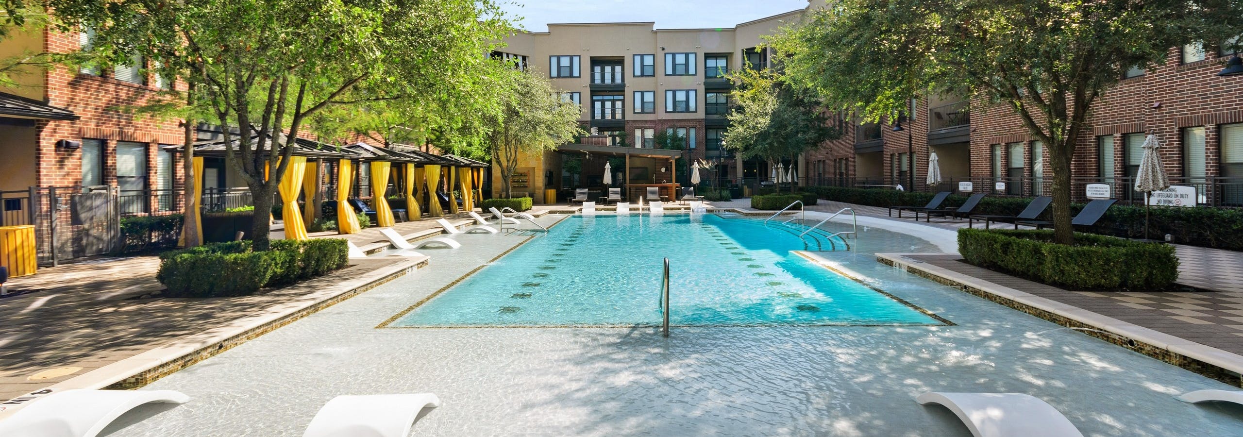 Courtyard pool at AMLI West Plano apartments featuring in water seating and surrounded by lush landscaping and lounge seating
