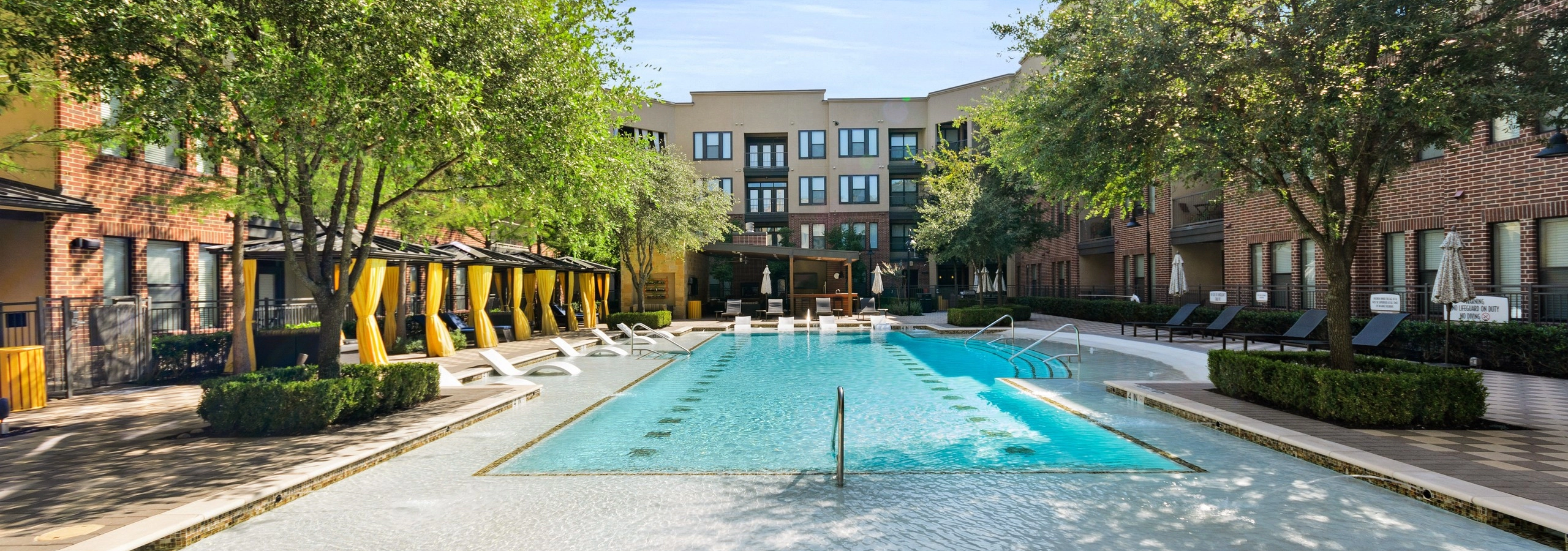 Courtyard pool at AMLI West Plano apartments featuring in water seating and surrounded by lush landscaping and lounge seating