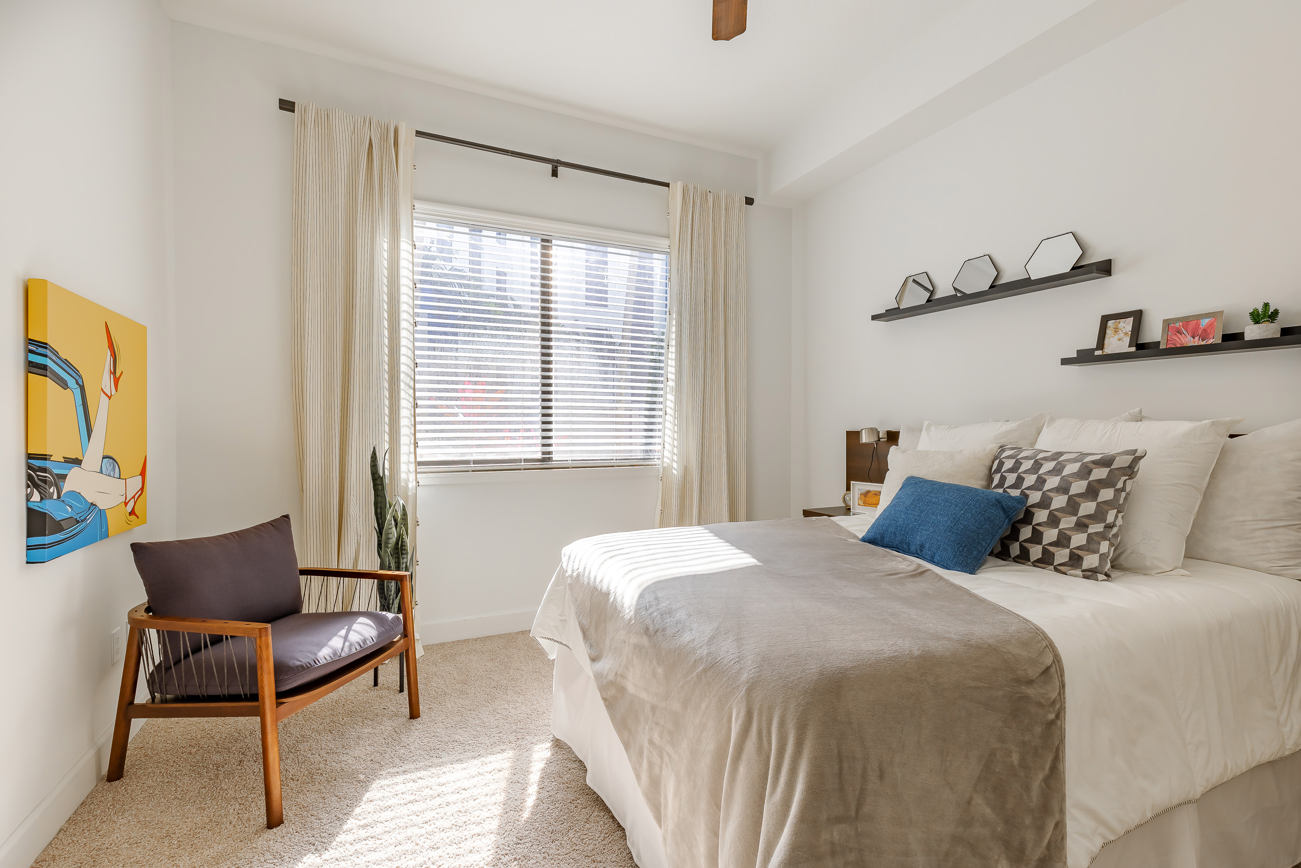 Bedroom at AMLI 8800 apartments with large window and bed with shelves above and bright yellow artwork and modern wood chair