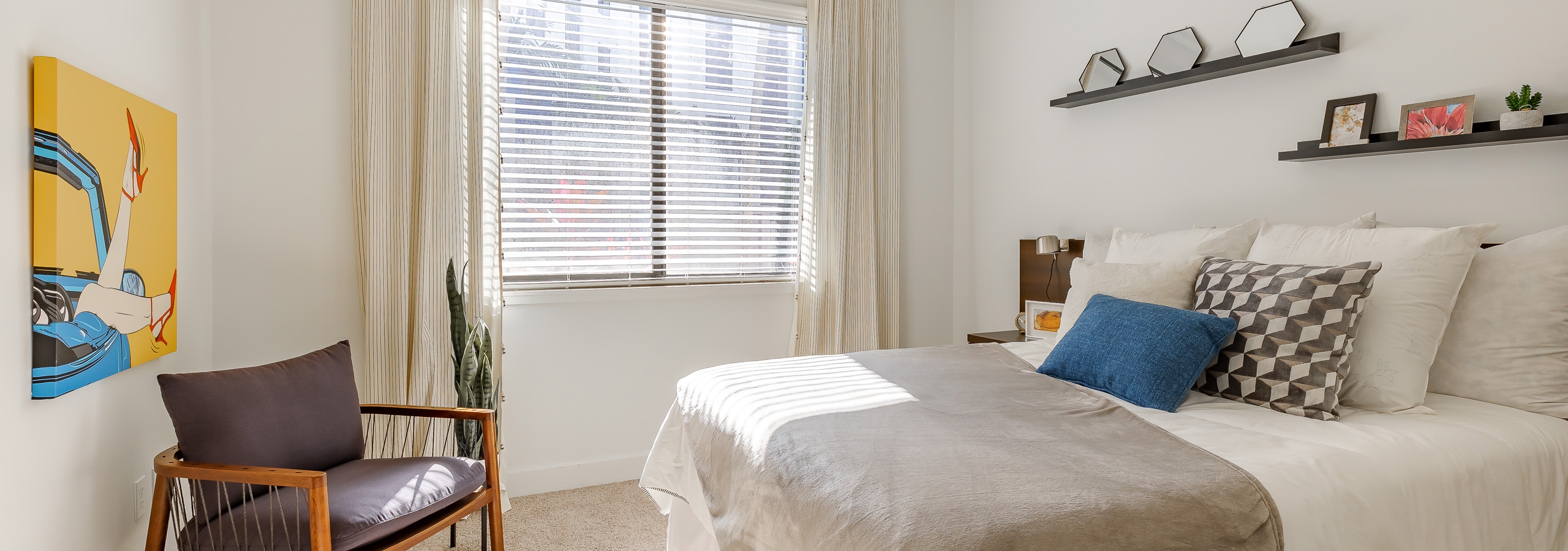 Bedroom at AMLI 8800 apartments with large window and bed with shelves above and bright yellow artwork and modern wood chair