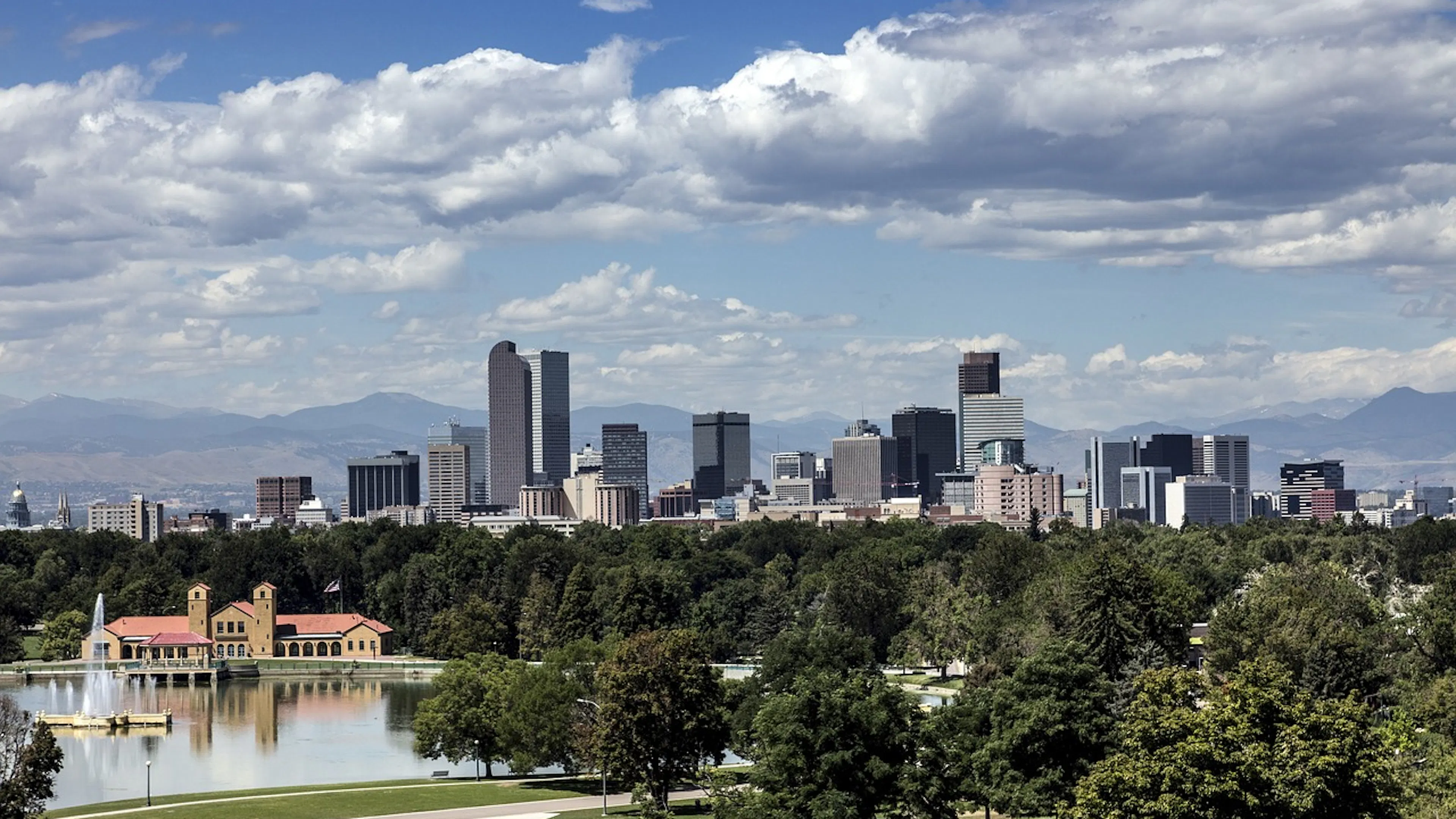 Denver skyline rising above green trees with a small lake in the foreground and a mountain range in the background