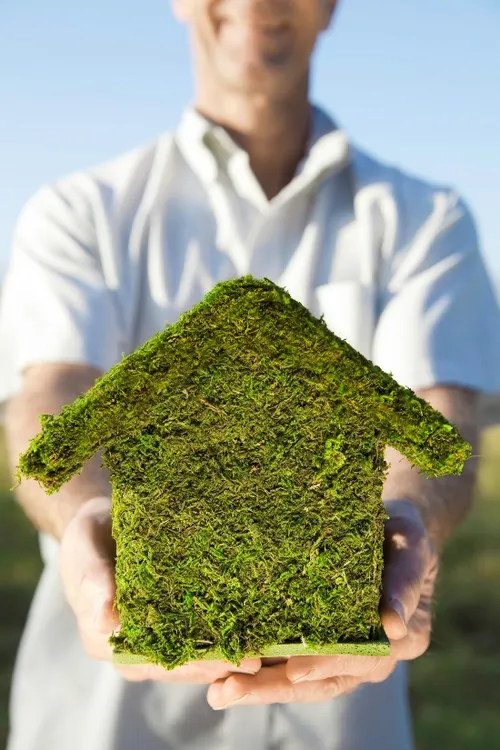A man in a white polo shirt holding a house figurine made of grass turf toward the camera