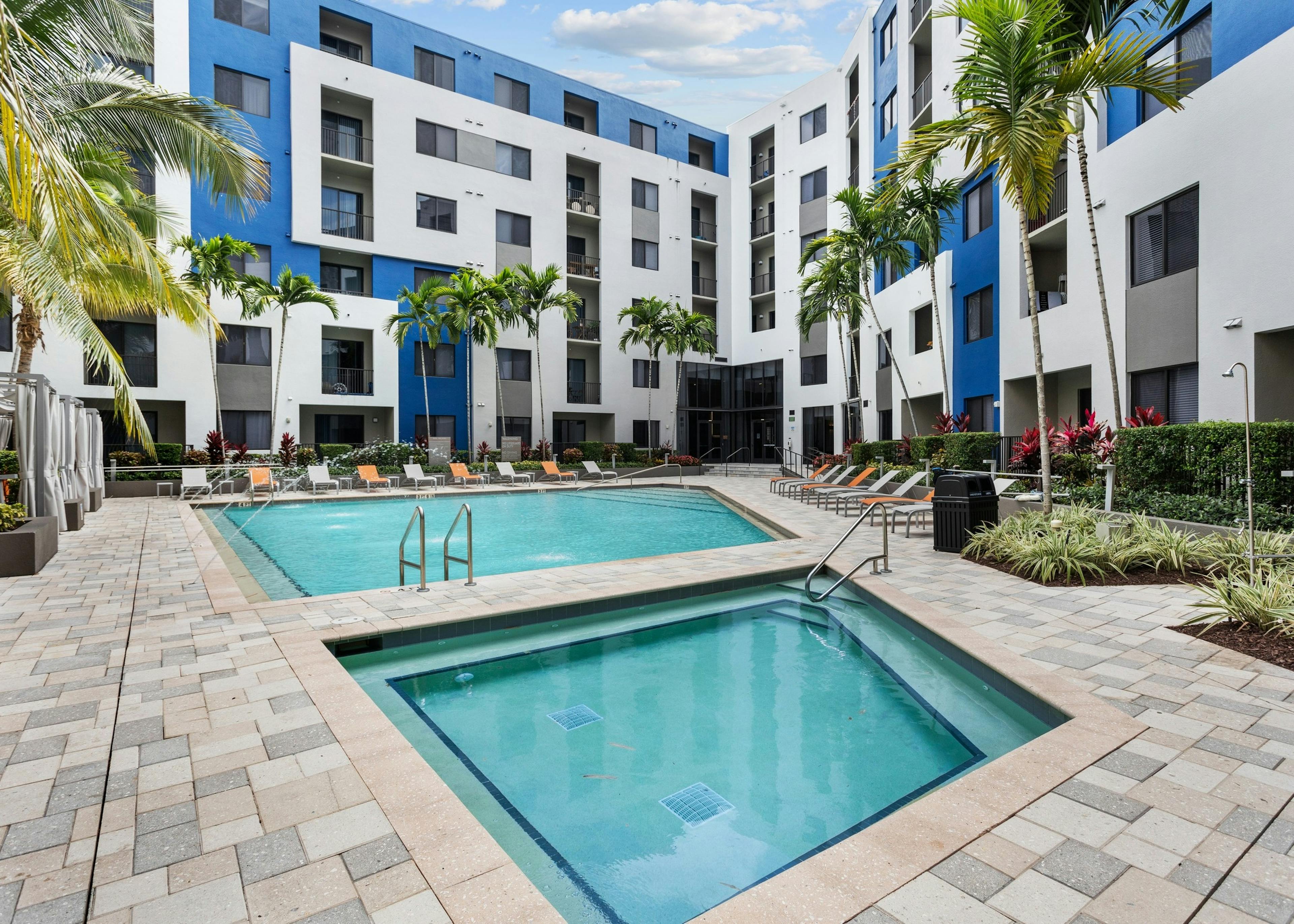 The pool at AMLI 8800 with water feature over pool and adjacent spa with gray and orange lounge chairs and cabanas 