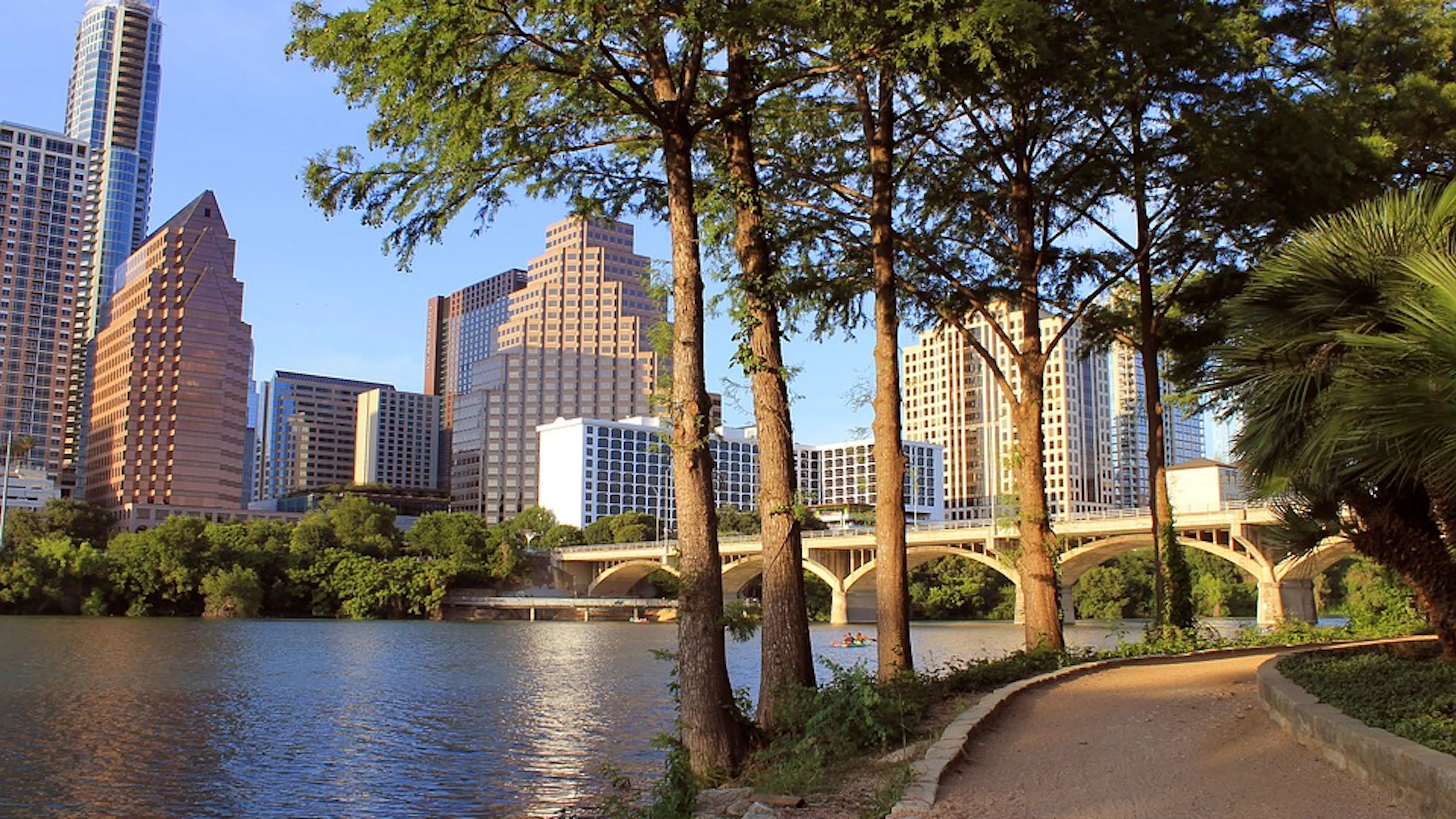 Lady Bird Lake flowing in front of the Downtown Austin skyline with a walking path in the foreground