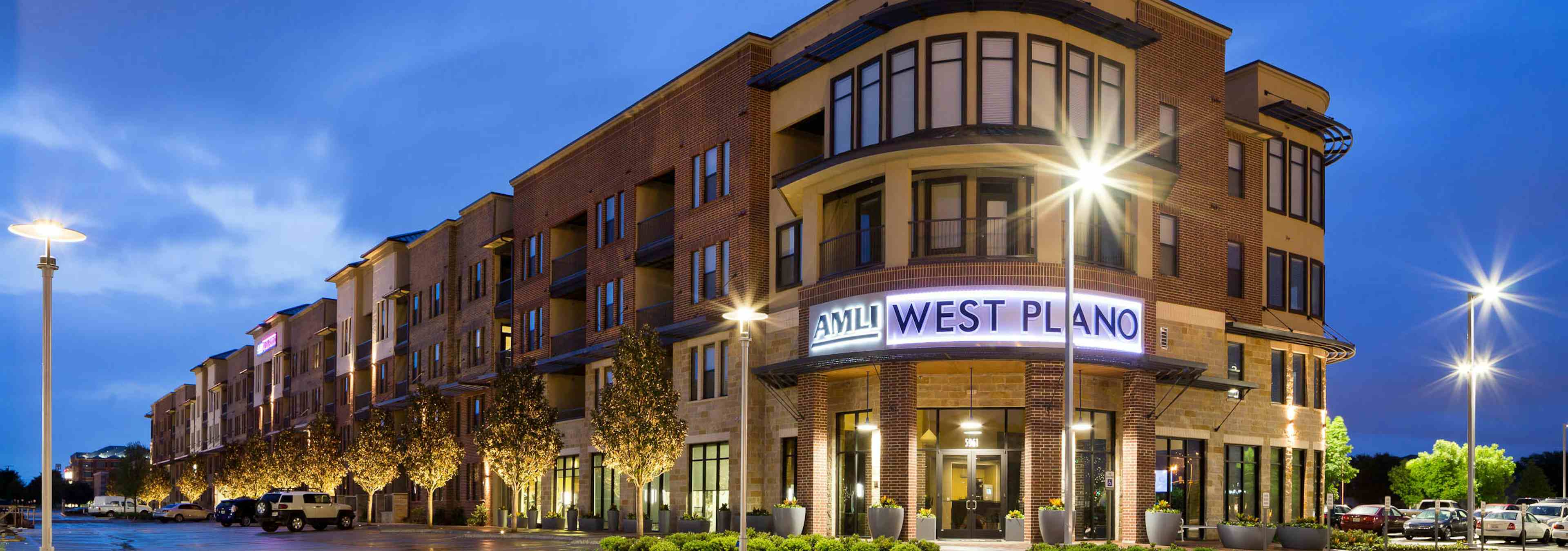 Exterior view of AMLI West Plano at nighttime with brick facade and lamp posts and trees neatly lining the building