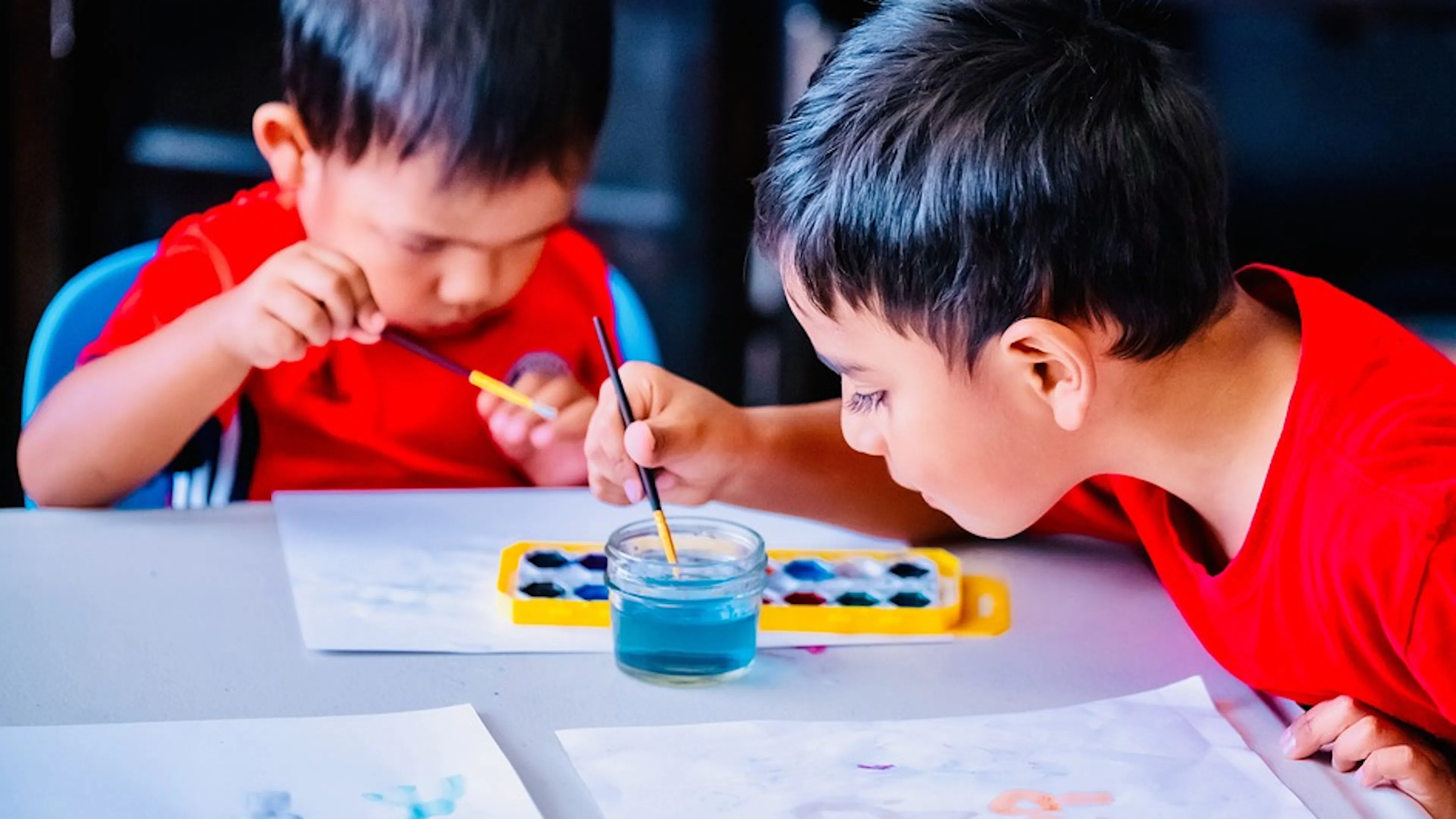 Two children wearing red t shirts hunched over a table with watercolor paints and paintbrushes