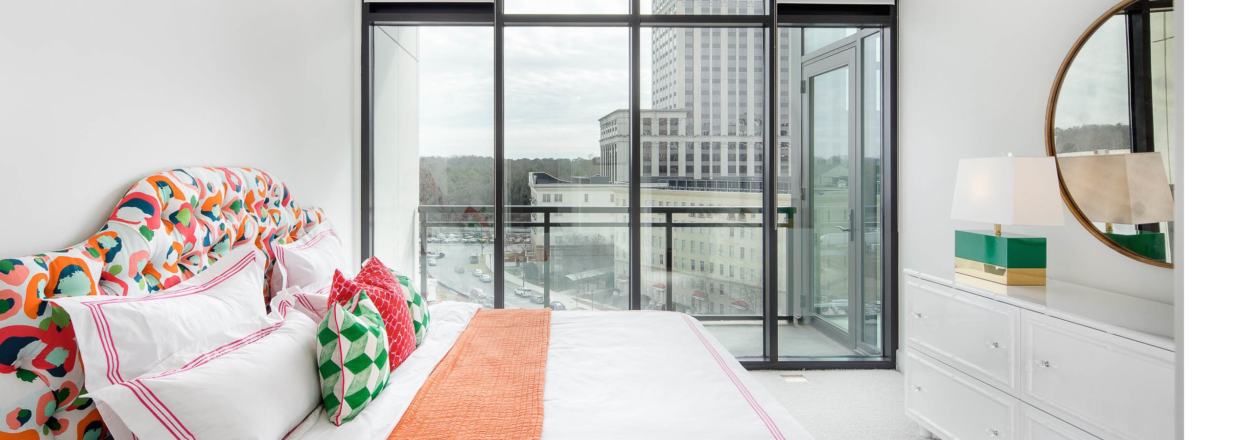 AMLI 3464 bedroom with floor to ceiling windows and a city view with white walls and bedding with bright coral and teal decor