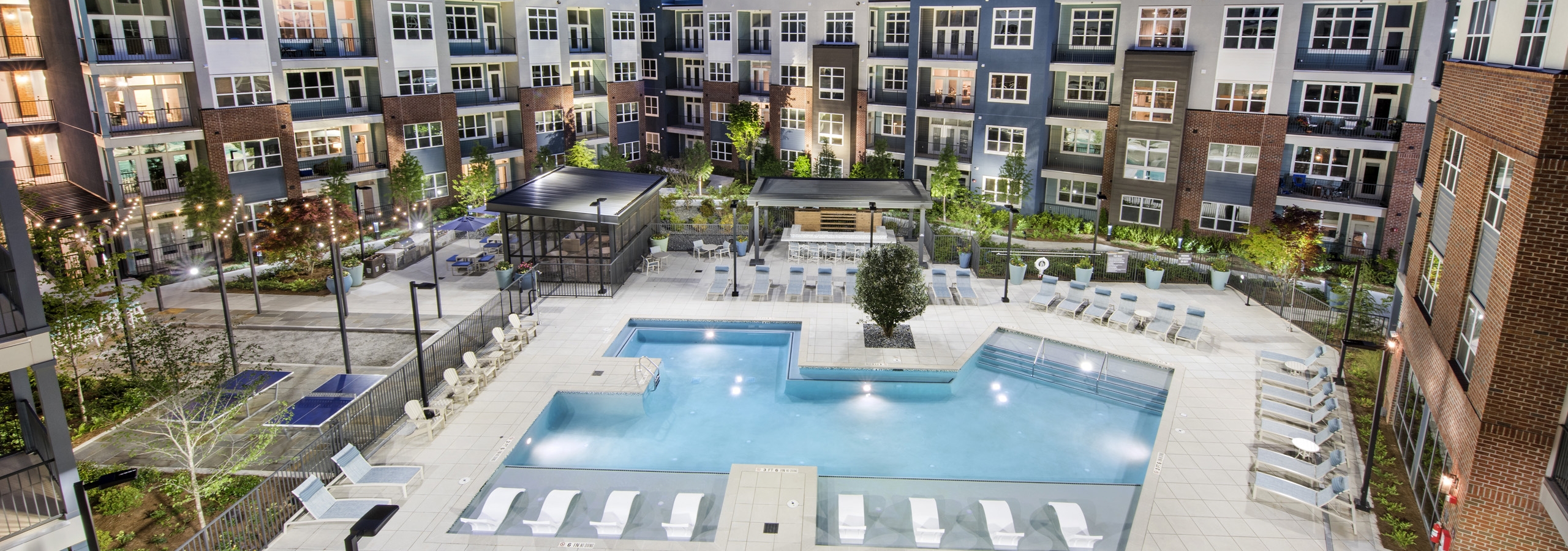 Overhead evening view of AMLI Decatur landscaped pool area with hanging lights and lounge chairs and uplit colorful building