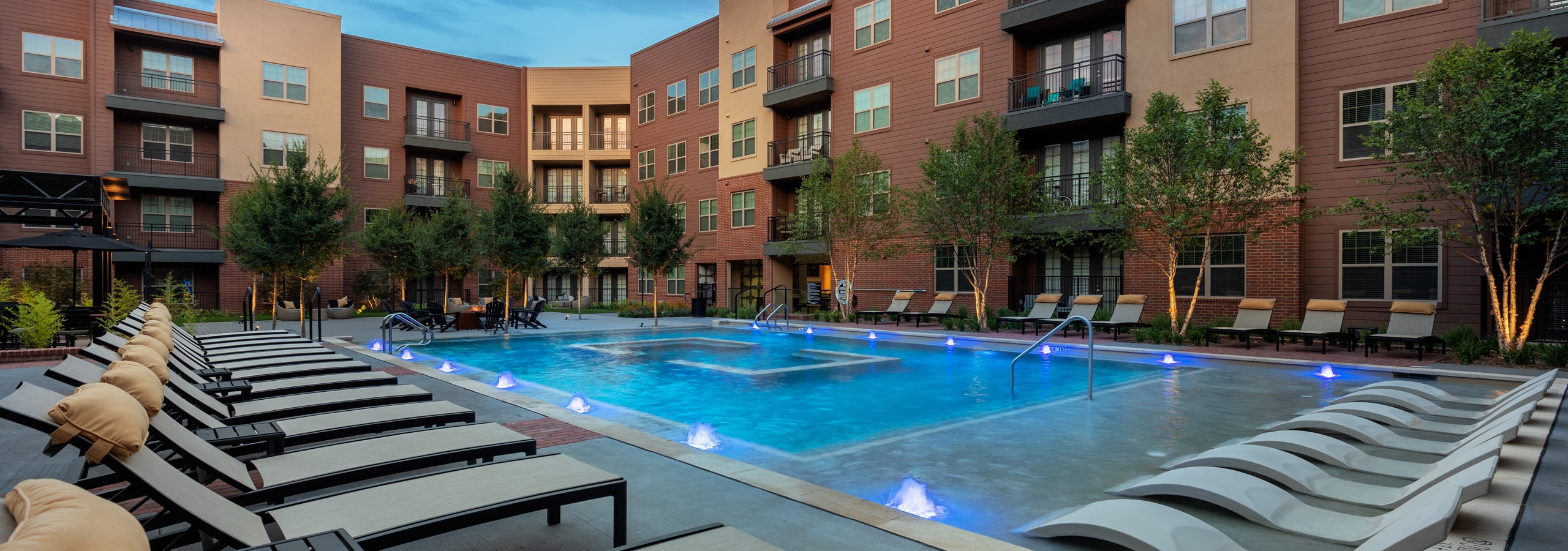 Evening view of AMLI Grapevine swimming pool with gray poolside lounges surrounded by brick building and green trees