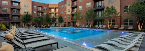 Evening view of AMLI Grapevine swimming pool with gray poolside lounges surrounded by brick building and green trees