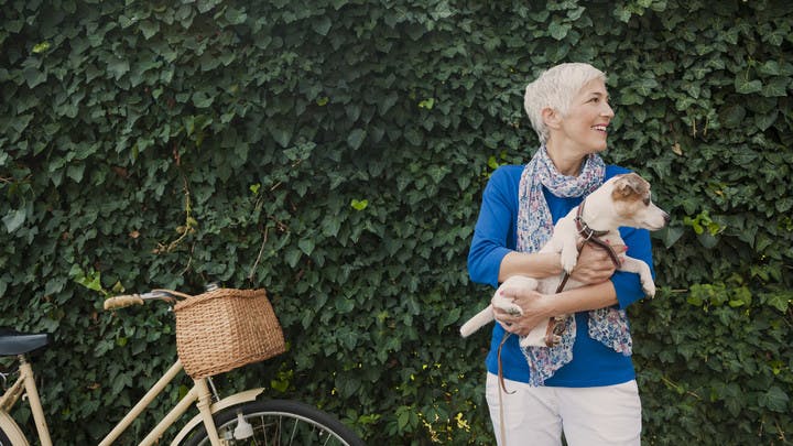Woman wearing a blue shirt standing against tall shrubbery holding a small dog and laughing with a bike standing to her left