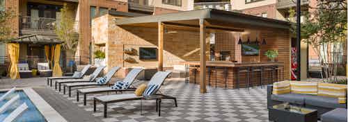 Outdoor kitchen in the AMLI West Plano pool area with barstools at a long counter and a view of surrounding chairs and trees
