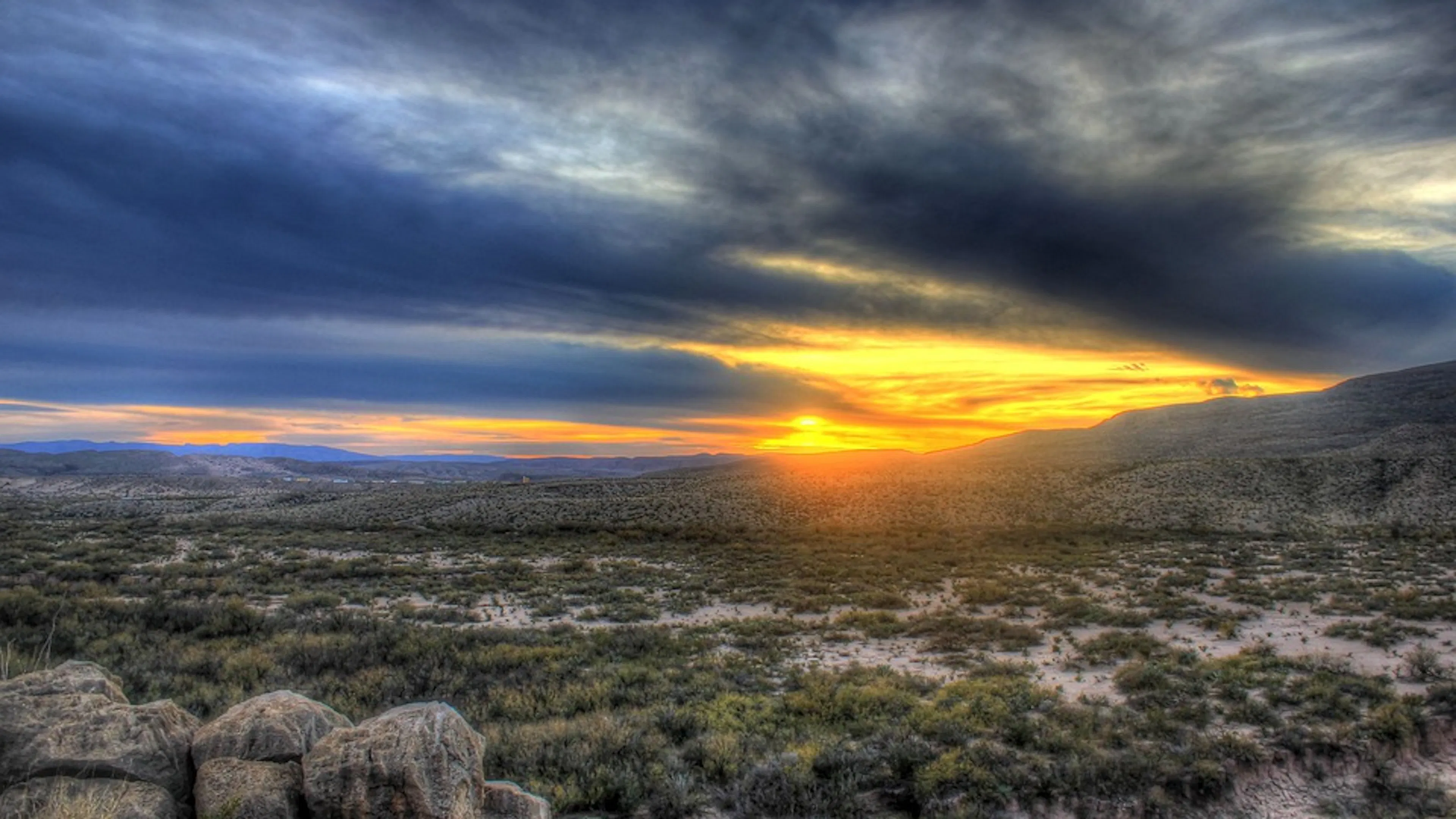 A vast Texas desert shrub landscape with boulders strewn on the sand and a sunset in the background