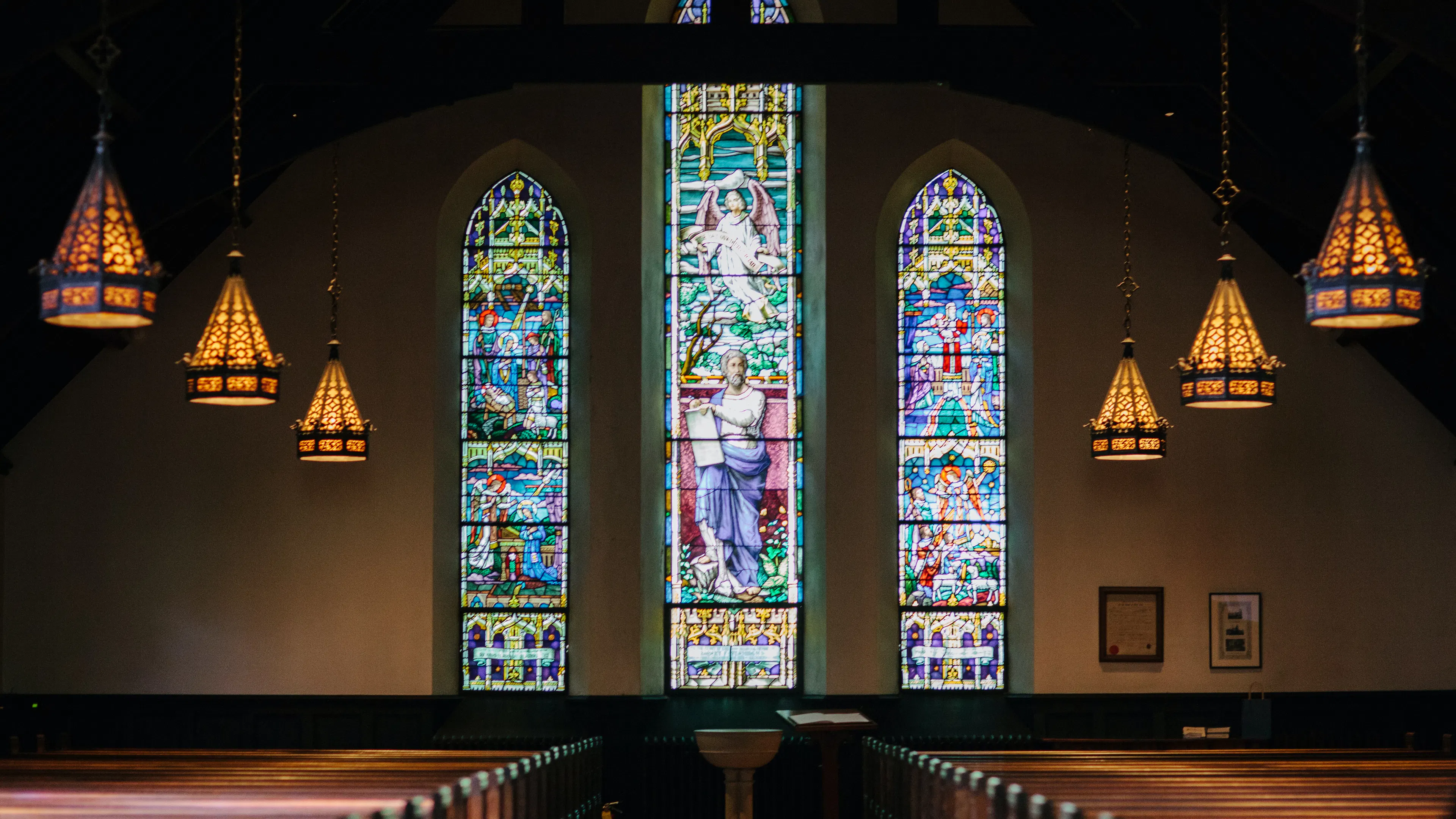 The interior of a church with three large stained glass windows shining light on the pews
