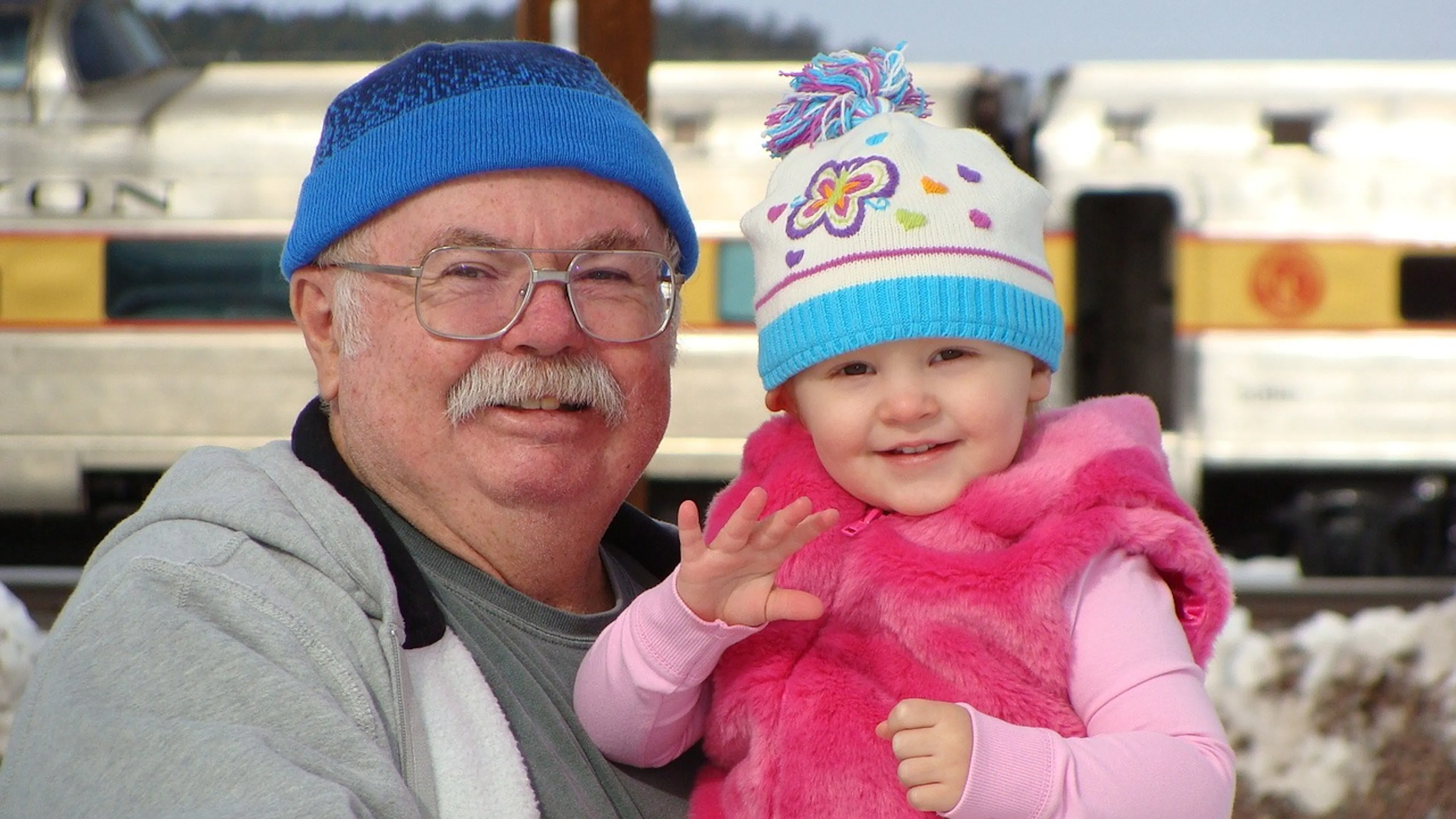 A grandpa wearing a blue hat and wide rim glasses holding a young girl in all pink and smiling for the camera