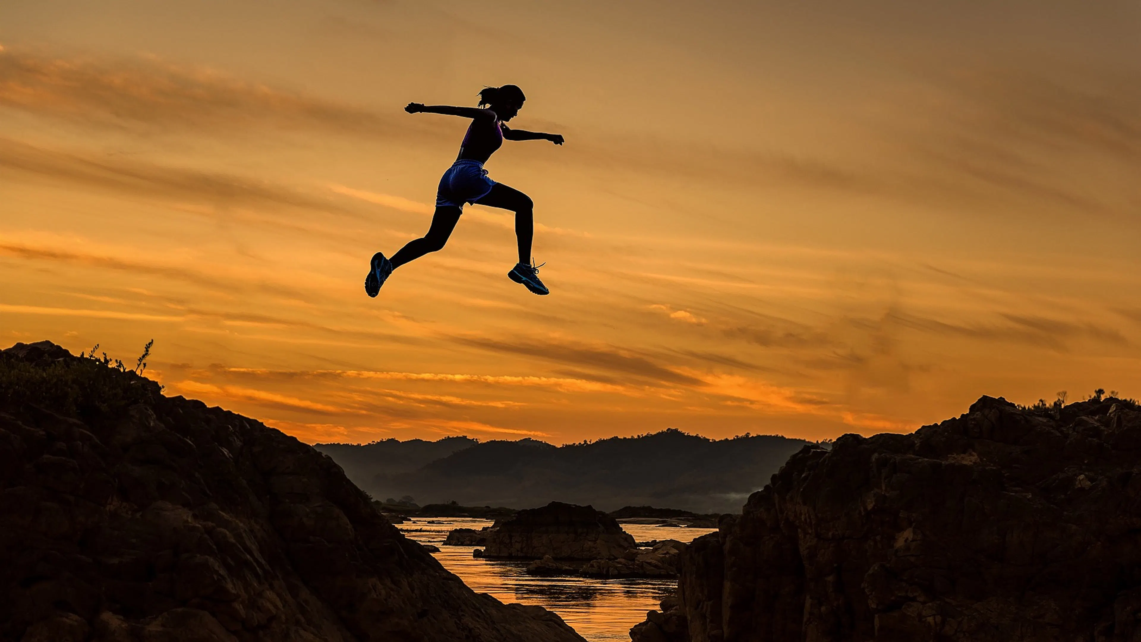 Silhouette of woman against an orange sky jumping over a body of water