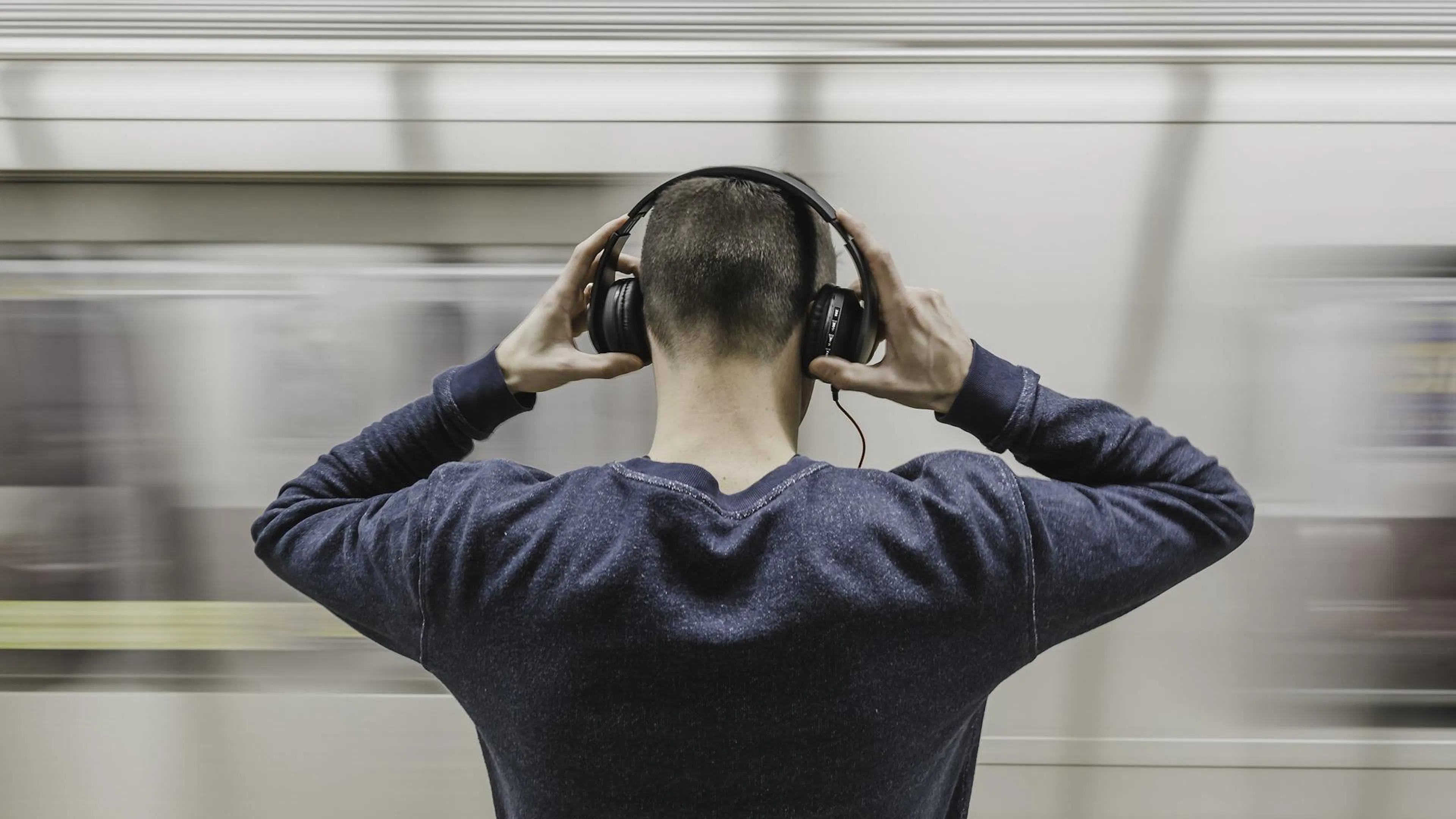 man standing in grey shirt with headphones on in front of moving subway train
