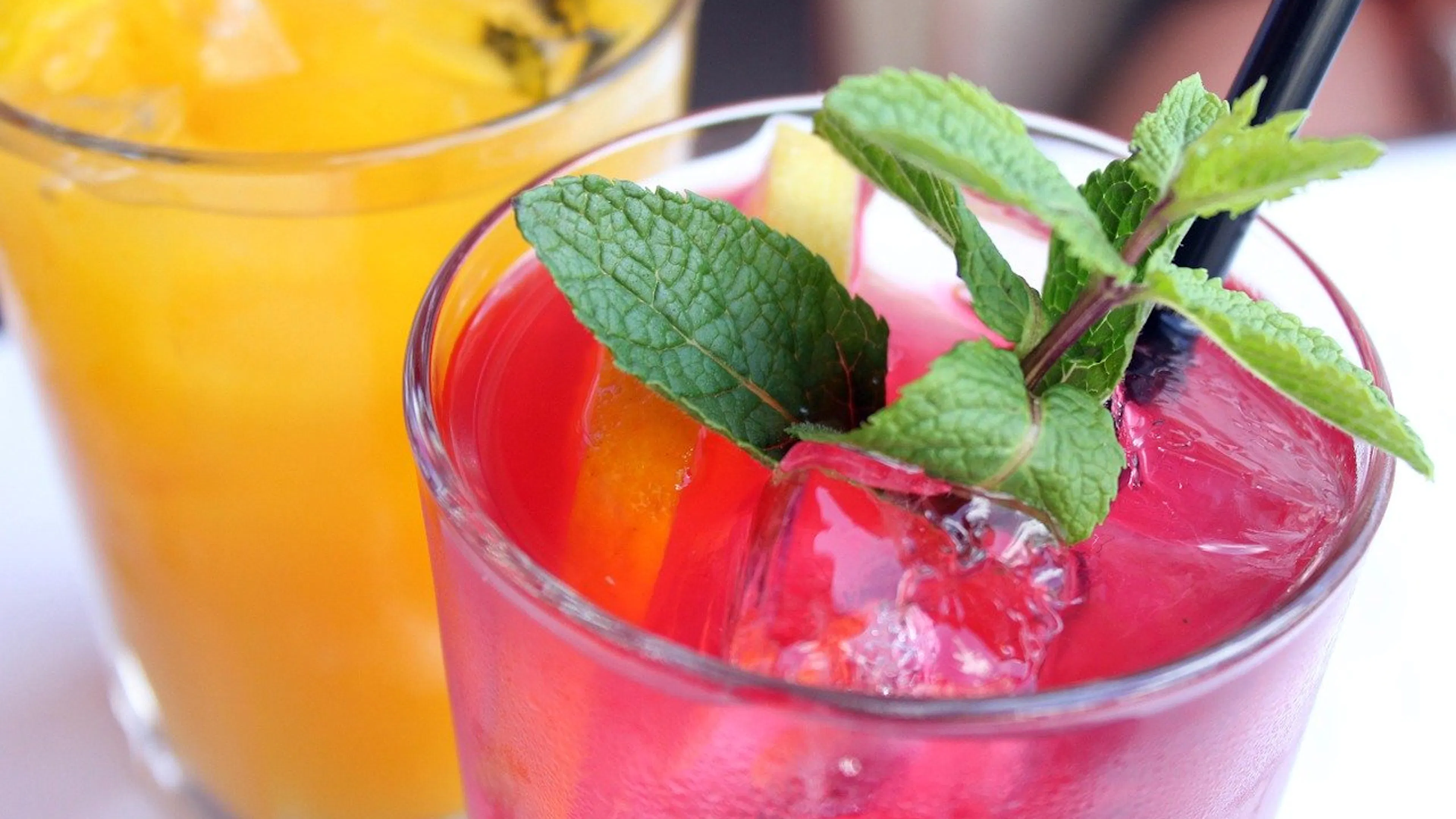 A pink cocktail in a pint glass that is garnished with sprigs of mint and a similar yellow cocktail behind it