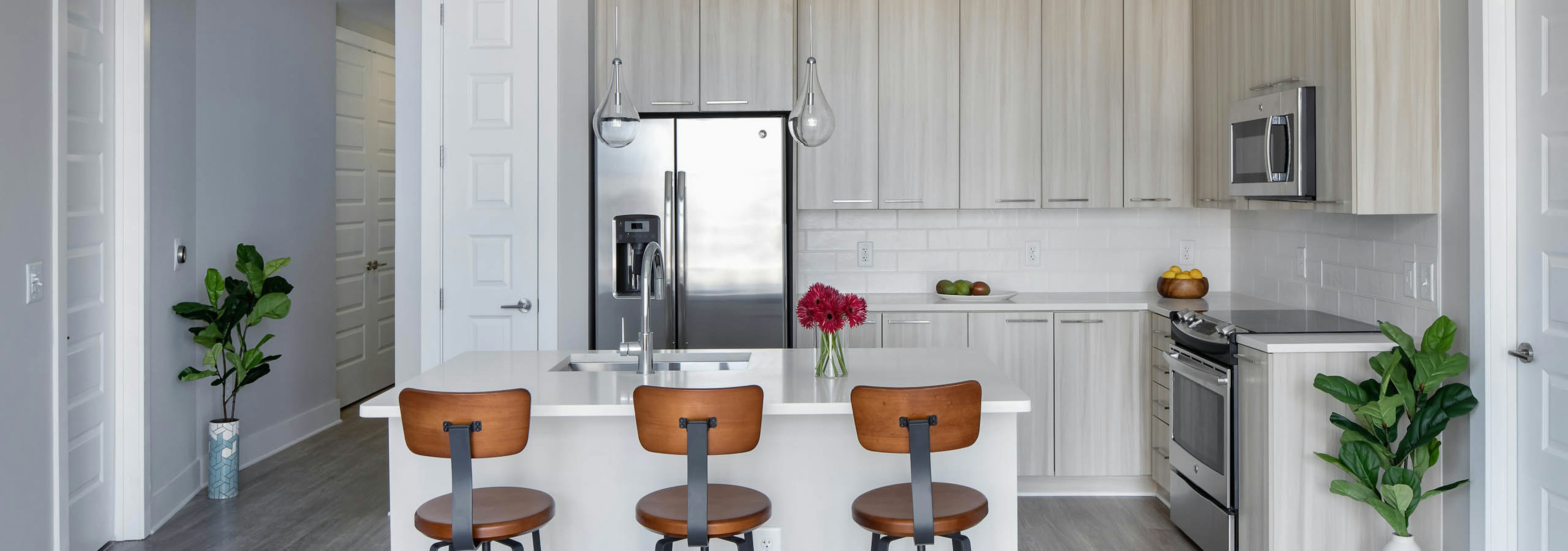 Kitchen at AMLI Arts Center with light wood cabinets and an island with three orange barstools and two faux floor plants