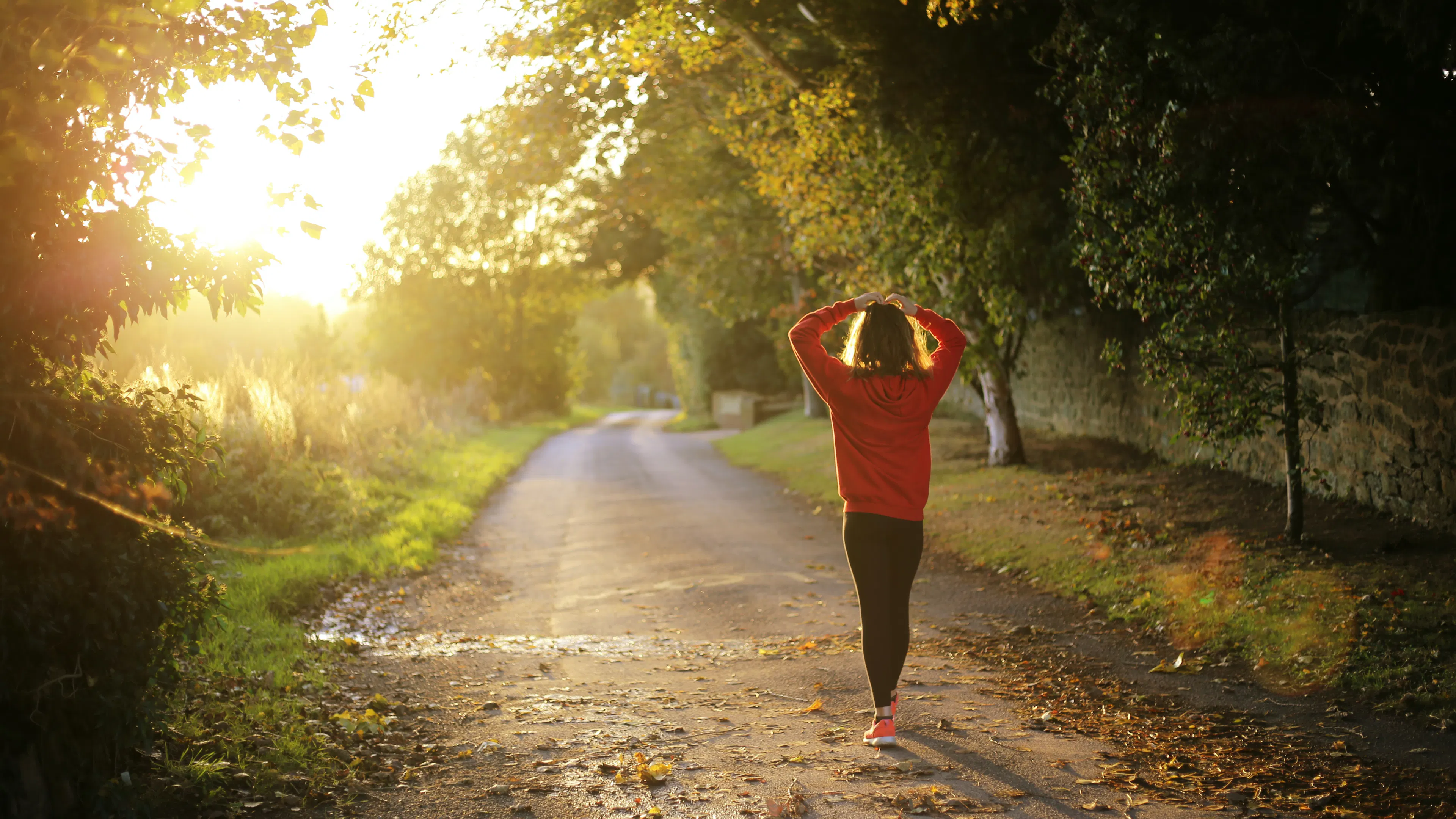 woman walking down a dirt road at sunrise in early fall with her hands on her head and leaves on the road
